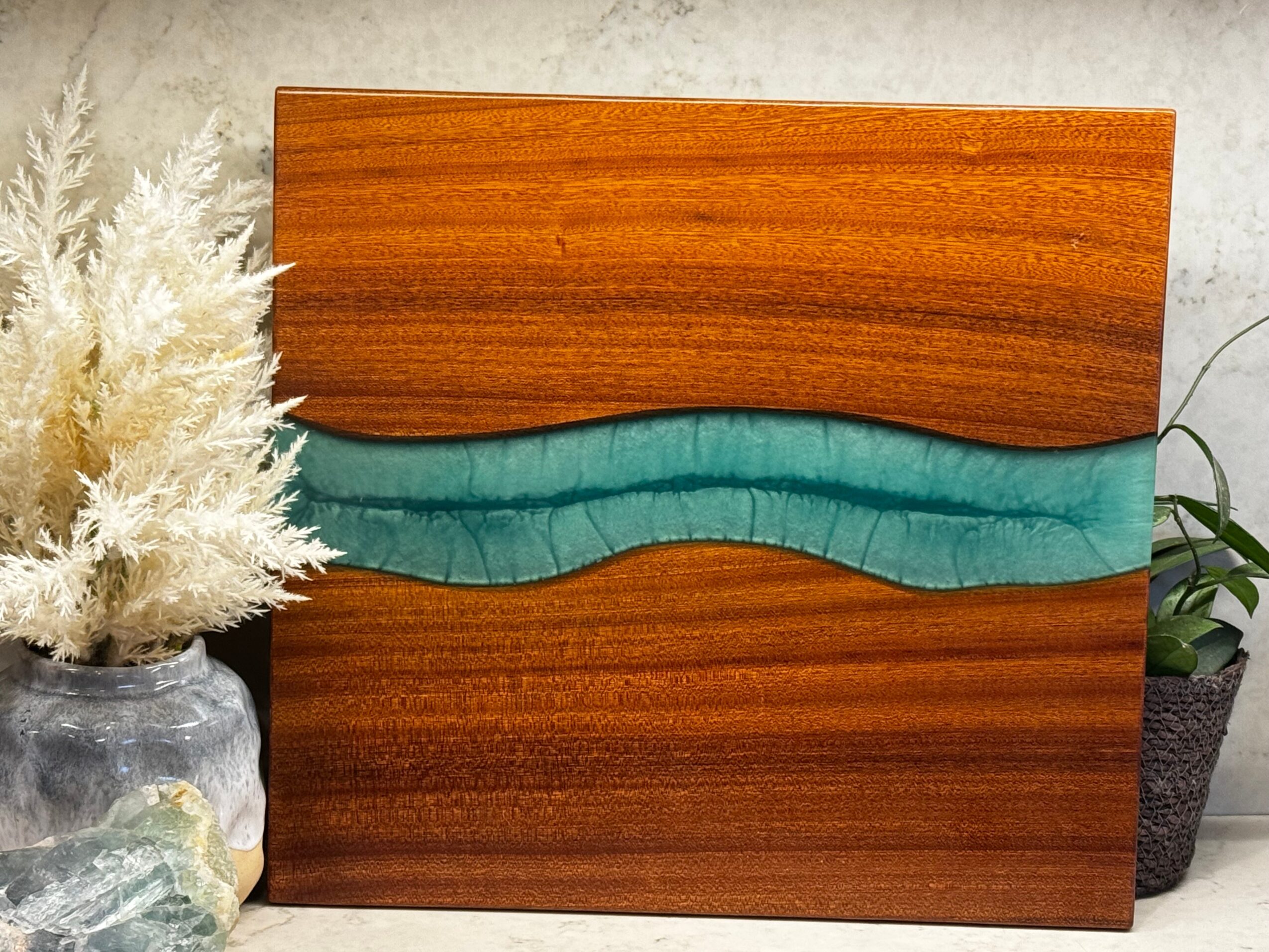 Decorative wooden panel with a wavy blue accent, flanked by potted plants and dried flowers, on a light background.