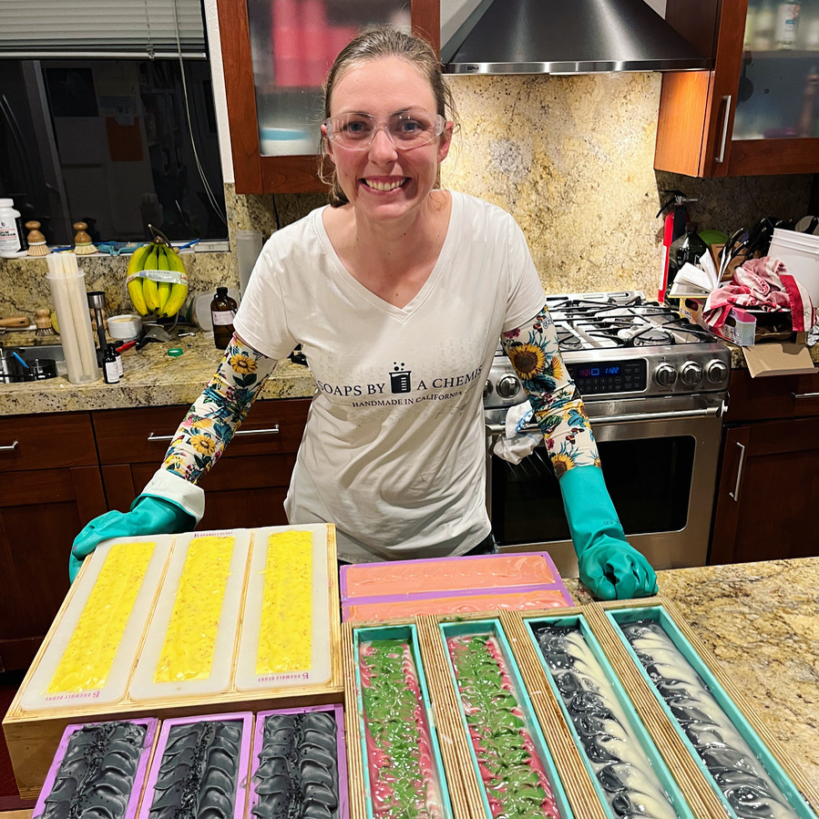 Woman smiling in a kitchen, surrounded by colorful soap molds on a countertop, wearing gloves and glasses.