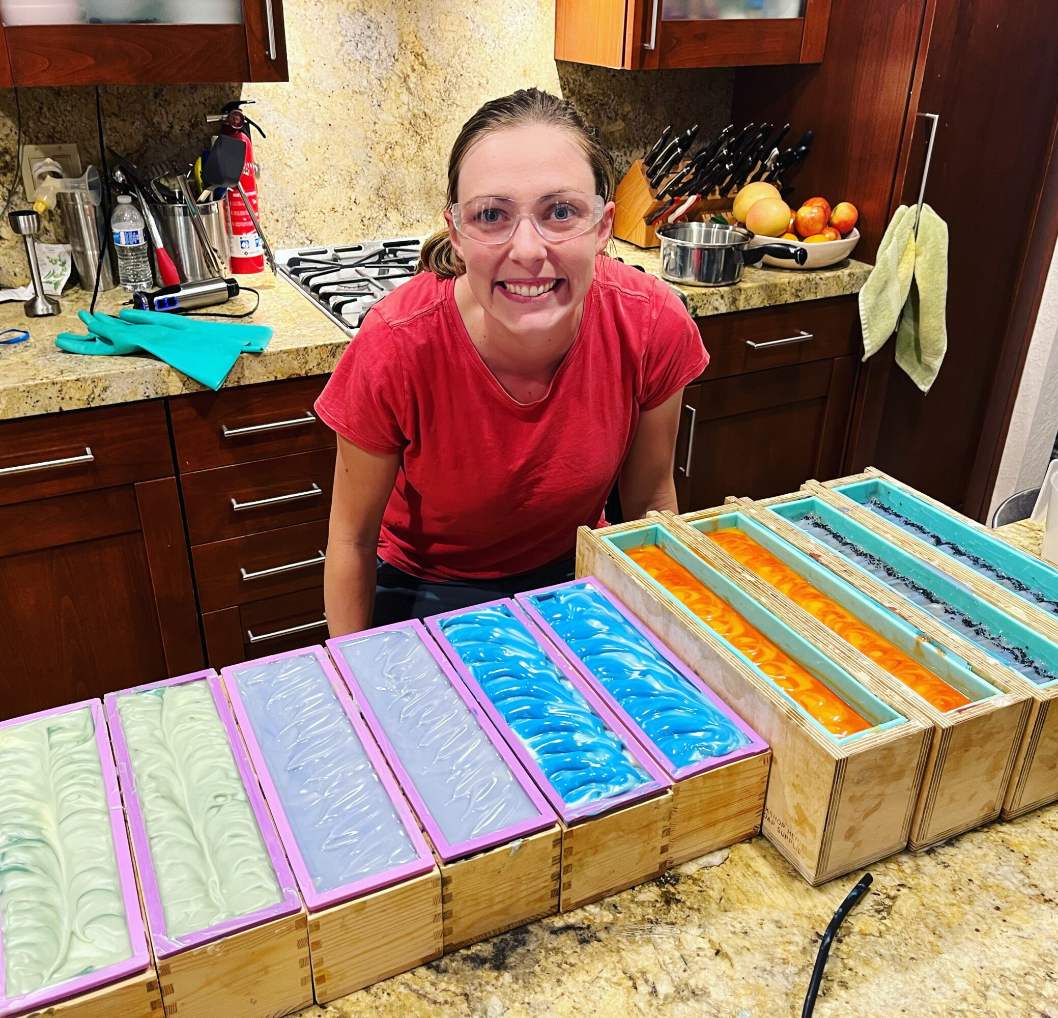 Smiling woman in red shirt standing behind a kitchen counter with colorful soap bars, wooden cabinets, and kitchen items.