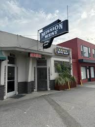 Building with a sign that reads 'MISSION WEST' and a smaller sign for a restaurant, with a potted plant outside.