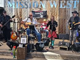 Band performs on stage with four musicians, one with guitar, in front of wooden backdrop, with flowers and equipment in foreground.