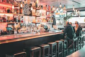 Bar with shelves filled with bottles and glasses, two people sitting on bar stools, illuminated by hanging lights.