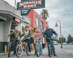 Four people with bicycles standing in front of a building with a sign that reads 'Mission West' under a cloudy sky.