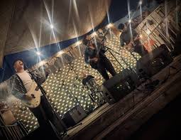 Group of people performing on stage with bright lights and decorated backdrop, some holding microphones.