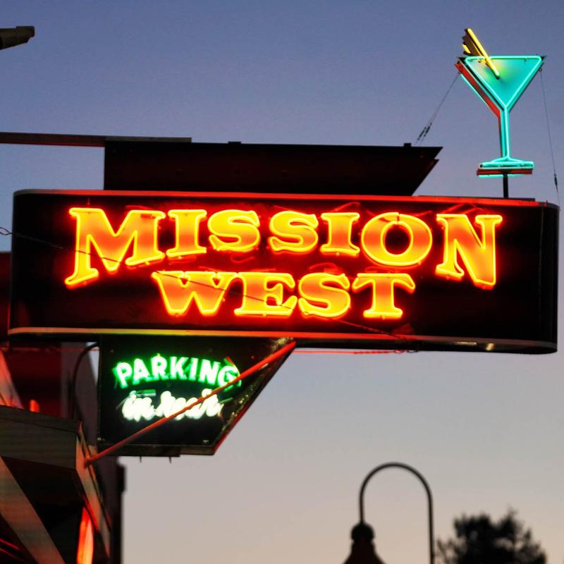 Neon sign for Mission West with a martini glass on top, and a parking sign below, against a dusk sky.