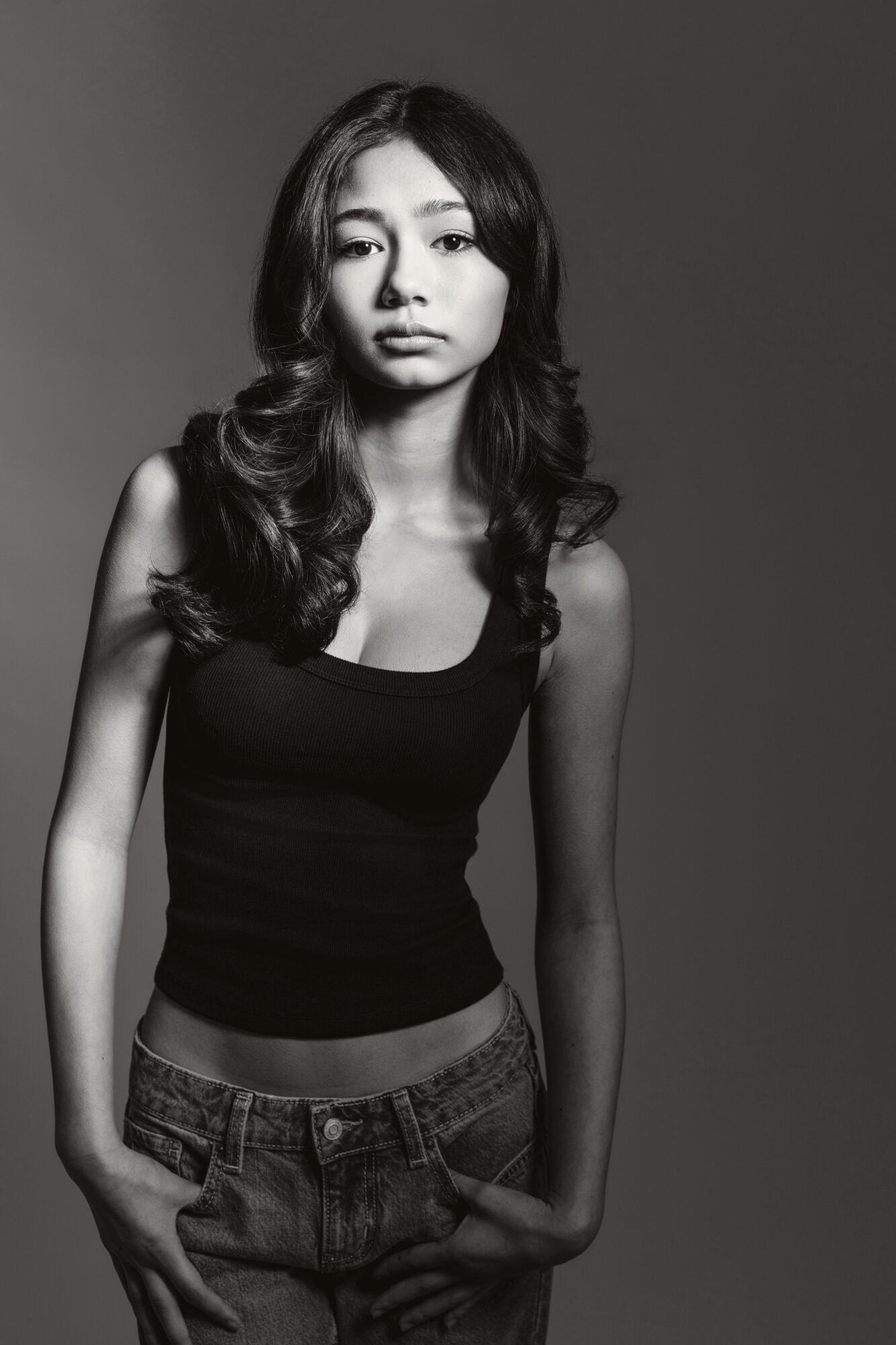 Young woman with long wavy hair wearing a sleeveless top and jeans, standing against a plain background.