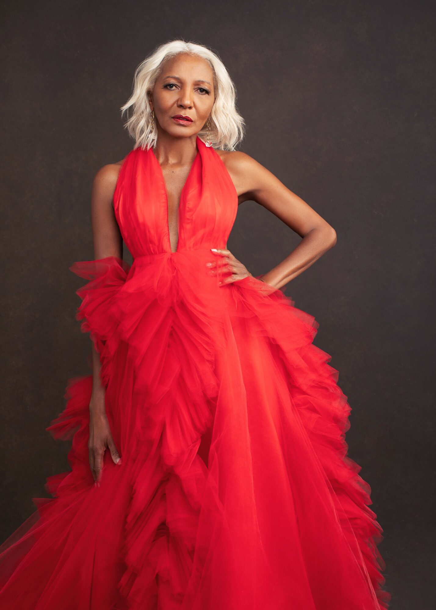 Woman with short white hair in a red gown with ruffles and a deep V neckline, standing against a dark background.