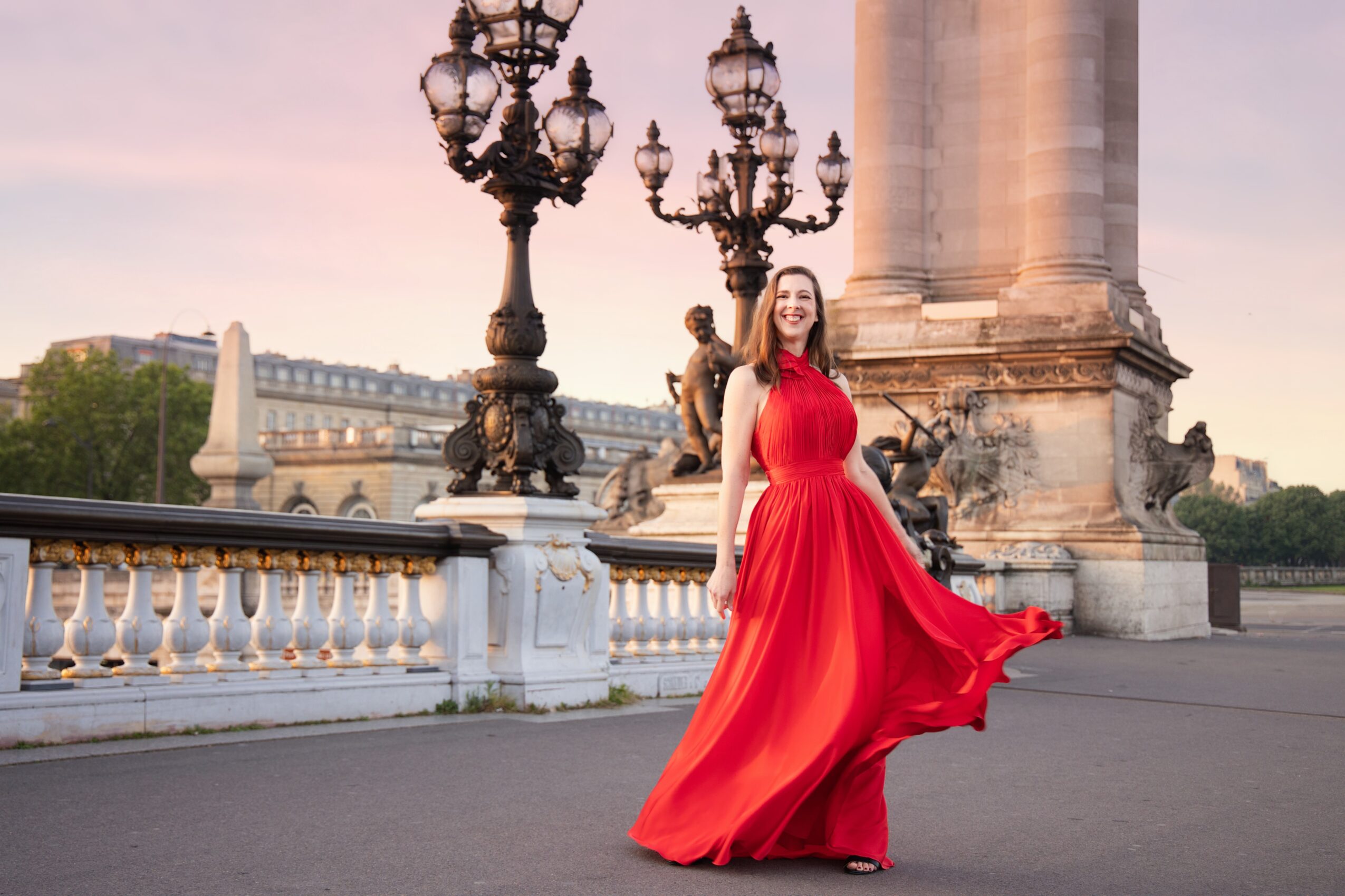 Woman in a flowing red dress standing near ornate street lamps and a stone railing, with a cityscape in the background.