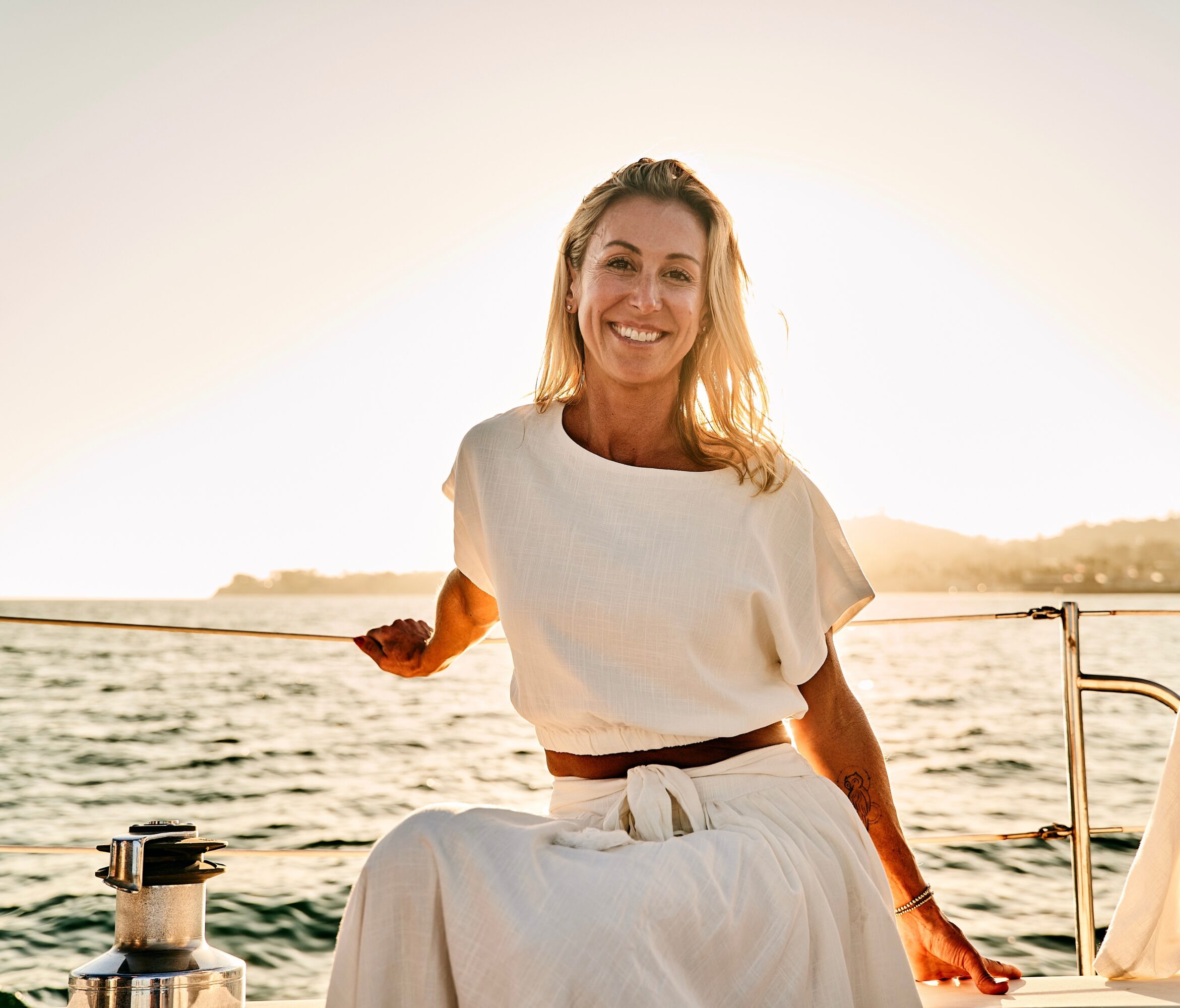 Woman smiling on a boat at sunset, holding a rope, wearing a white dress, water and distant land in background.