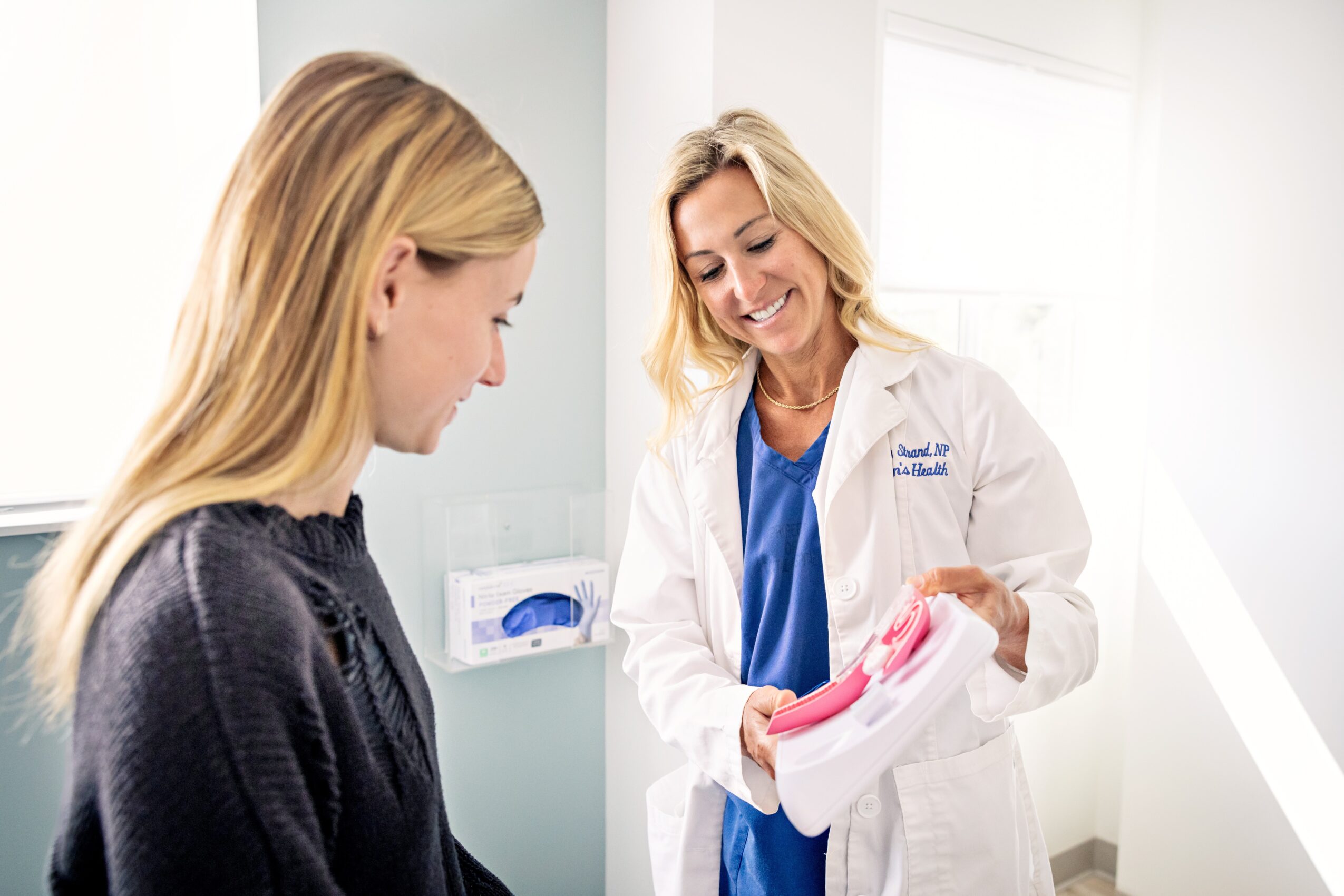 A female doctor and a young woman are in a bright medical office, smiling and talking, with the doctor holding a pink object.