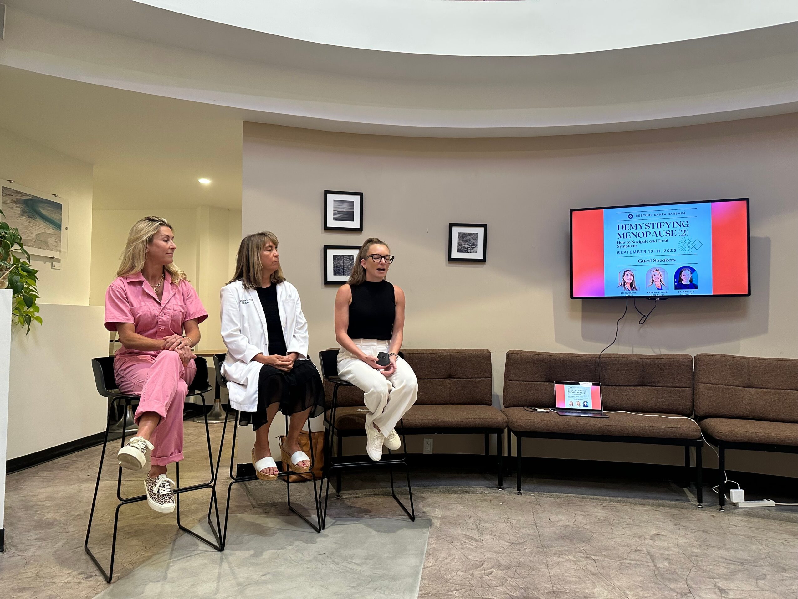 Three women sit on high stools in a room with a curved ceiling, a wall-mounted screen, and a long sofa.