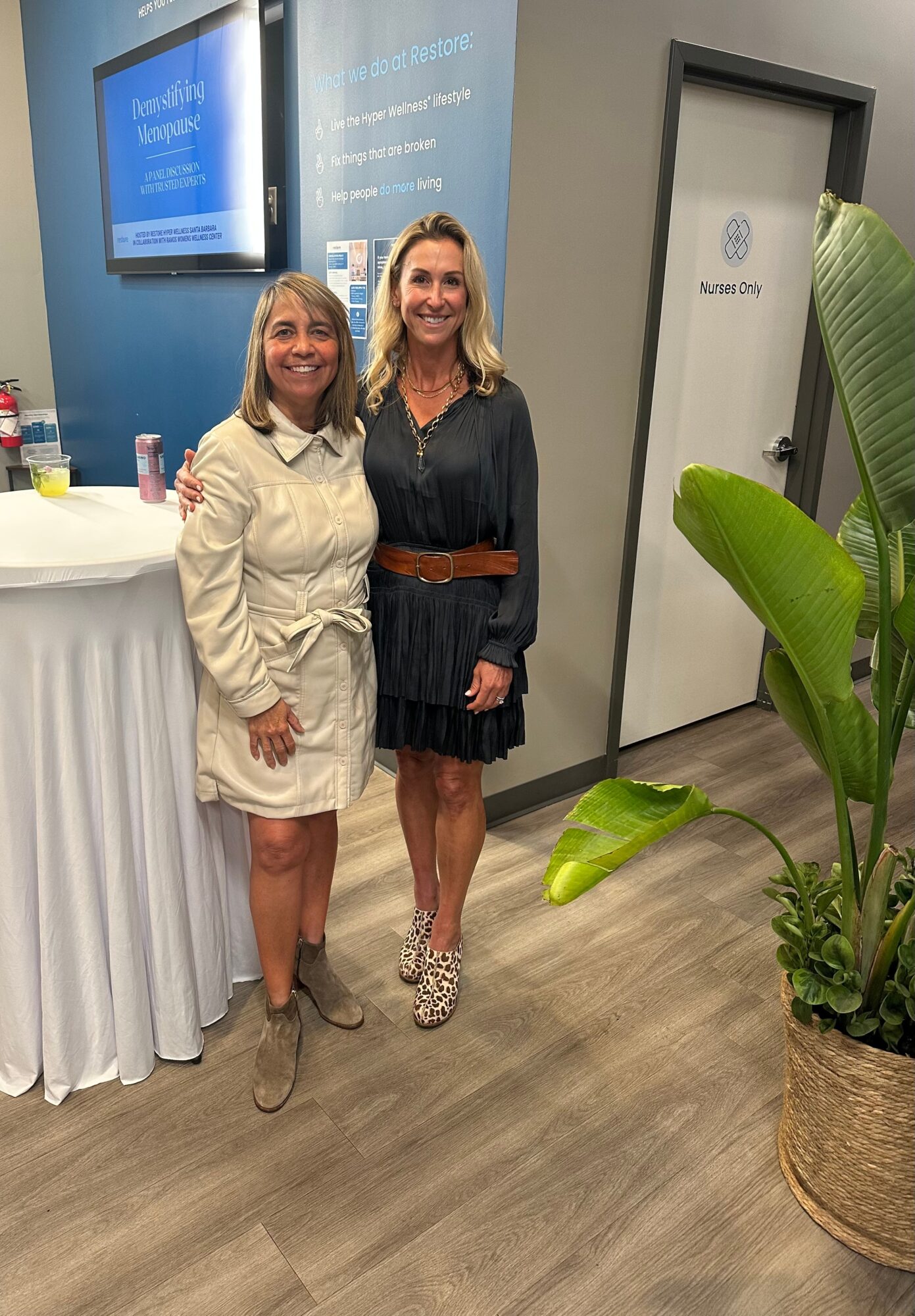 Two women stand indoors near a potted plant and a door, smiling at the camera, with a blue display screen behind them.