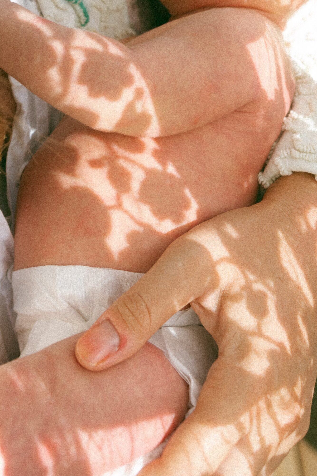 Close-up of a baby's body with shadows of leaves on skin, lying on a white surface, wearing a diaper.