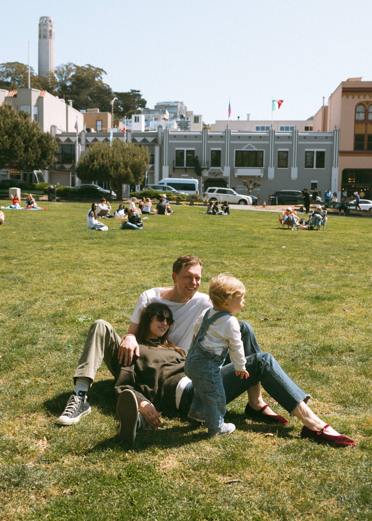 Three people sitting on grass in a park, with buildings and other people in the background.
