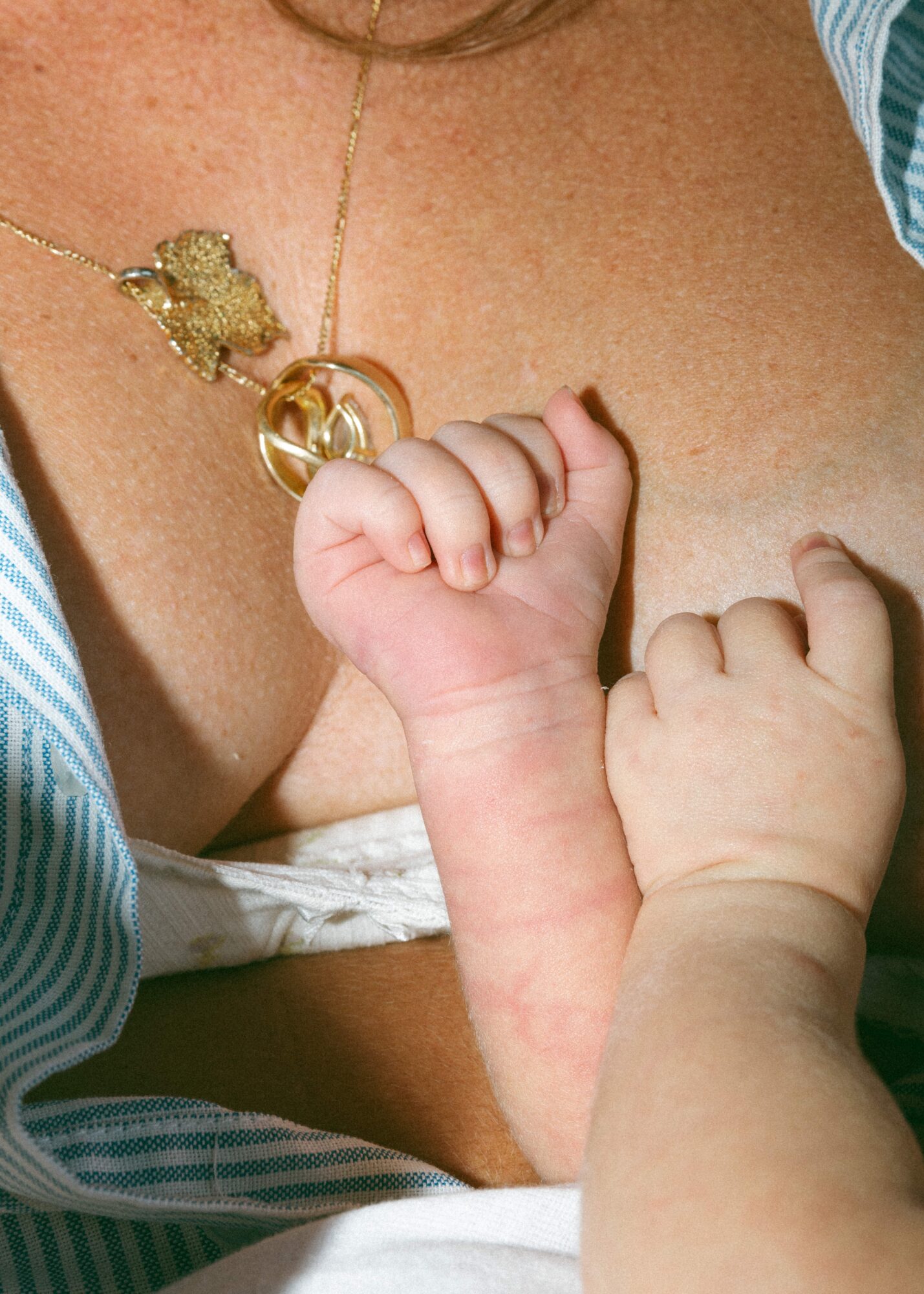 Close-up of a baby's hand gripping an adult's chest, with a necklace visible, skin tones, and clothing details.