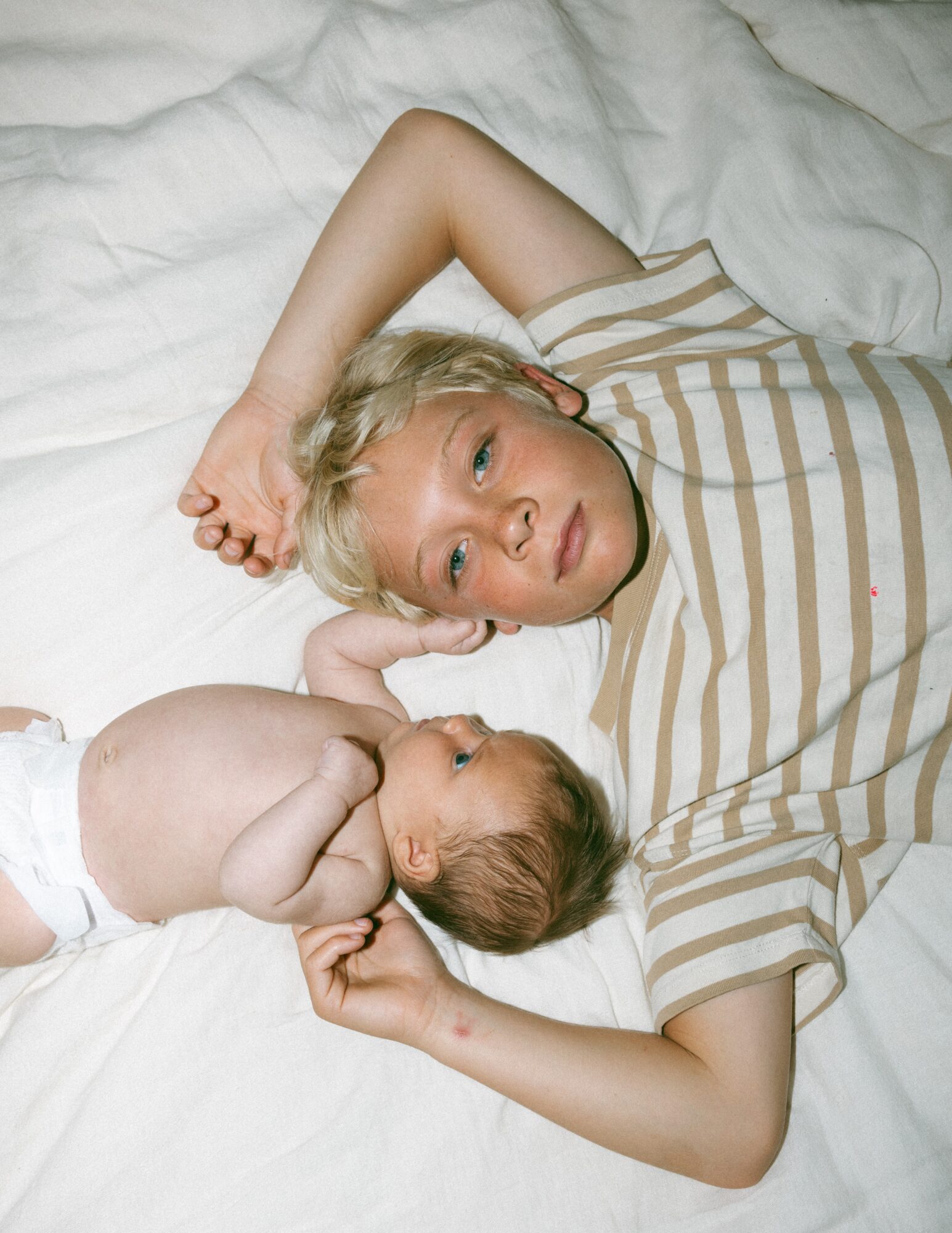 A young person with short blonde hair lying on a bed, looking at a baby with light brown hair, both on white sheets.