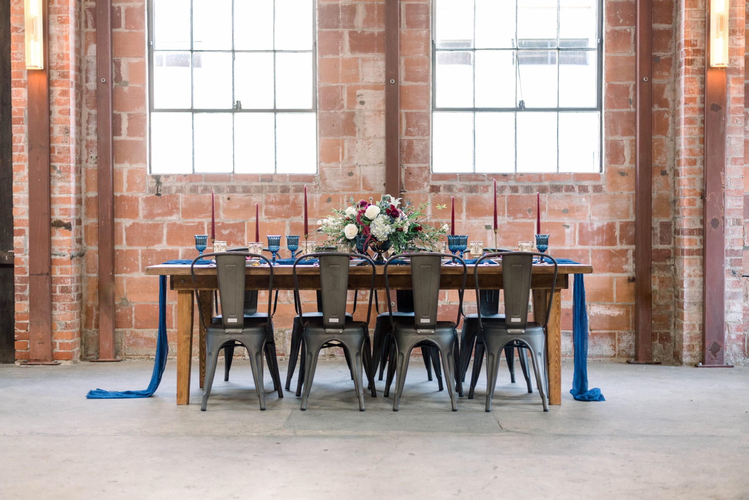 Long wooden table with eight black chairs, floral centerpiece, set with blue napkins, in an industrial-style brick room.