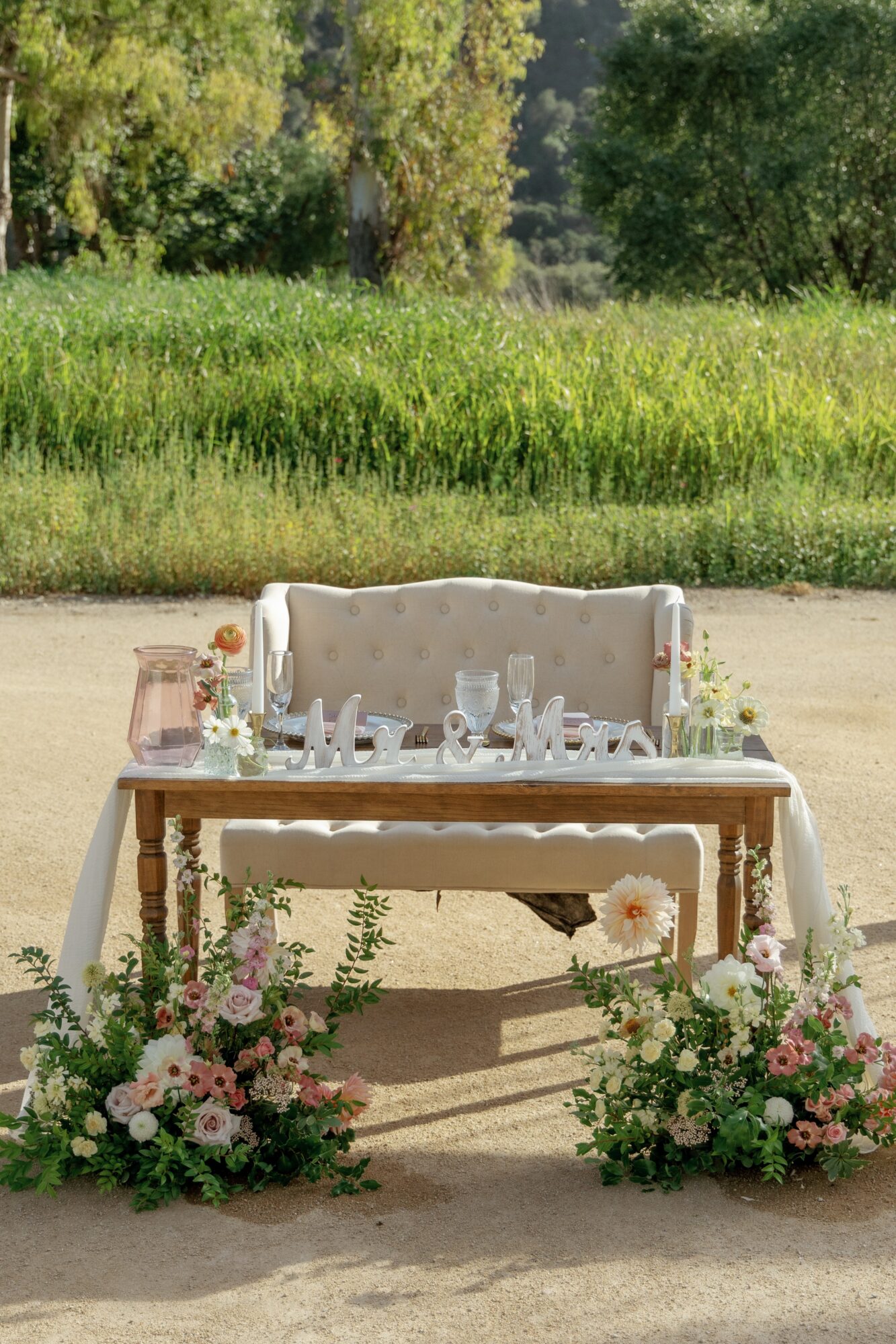 Table with floral arrangements and glassware outdoors, with green grass and trees in background.