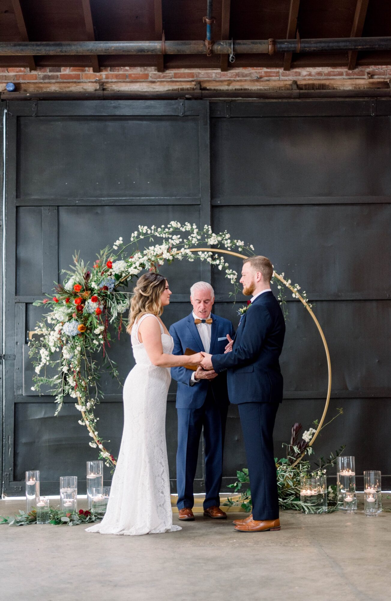 Bride and groom holding hands during wedding ceremony, officiant standing behind them, floral arch behind, candles on the floor.