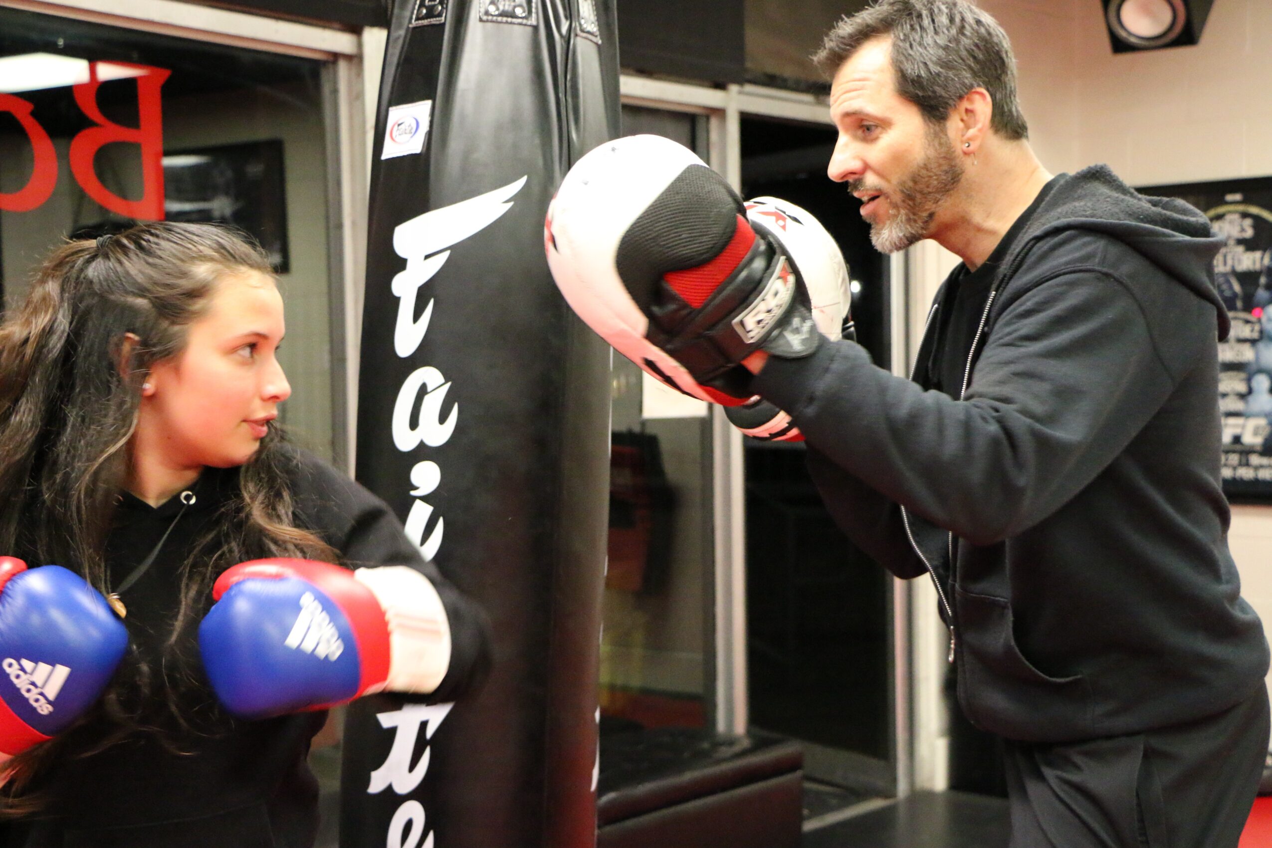 A man in black training gloves practices boxing with a young girl in a gym, focusing on a punching bag.