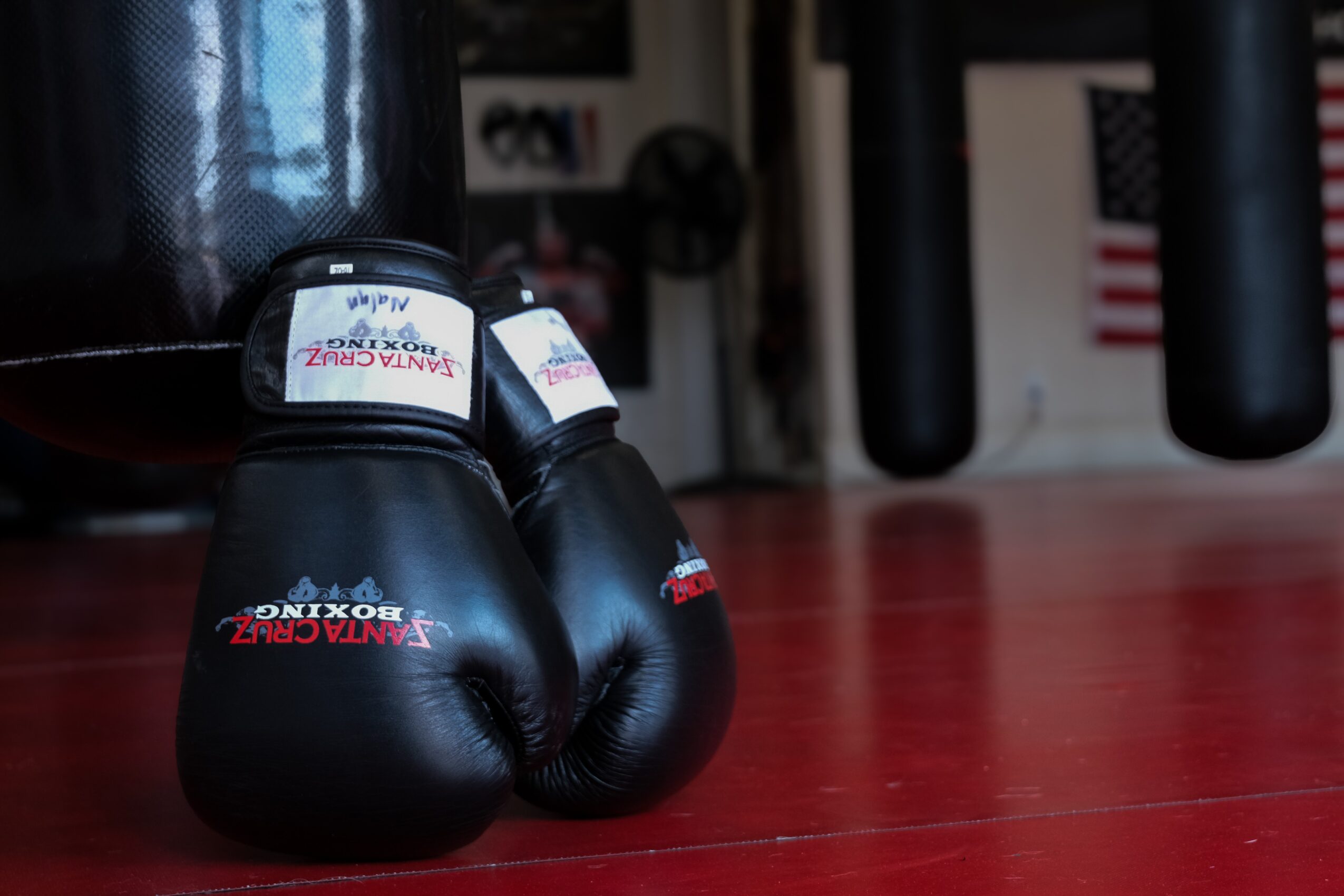 Pair of black boxing gloves on a red floor in a gym, with punching bags in the background.