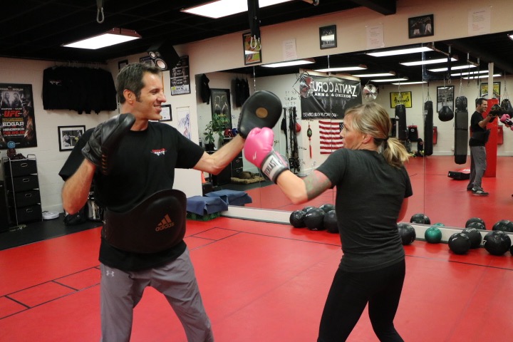 Two women boxing in a gym, one throwing a punch, the other blocking, with mirrors and equipment in the background.