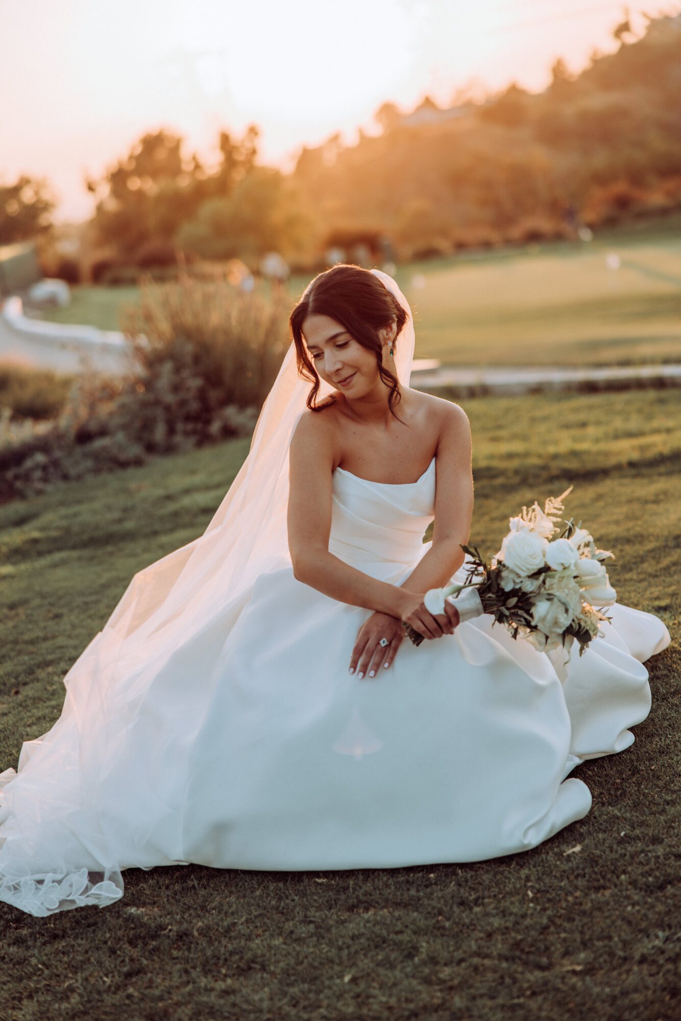 Bride in a white wedding dress sitting on grass with a bouquet, smiling outdoors during sunset.