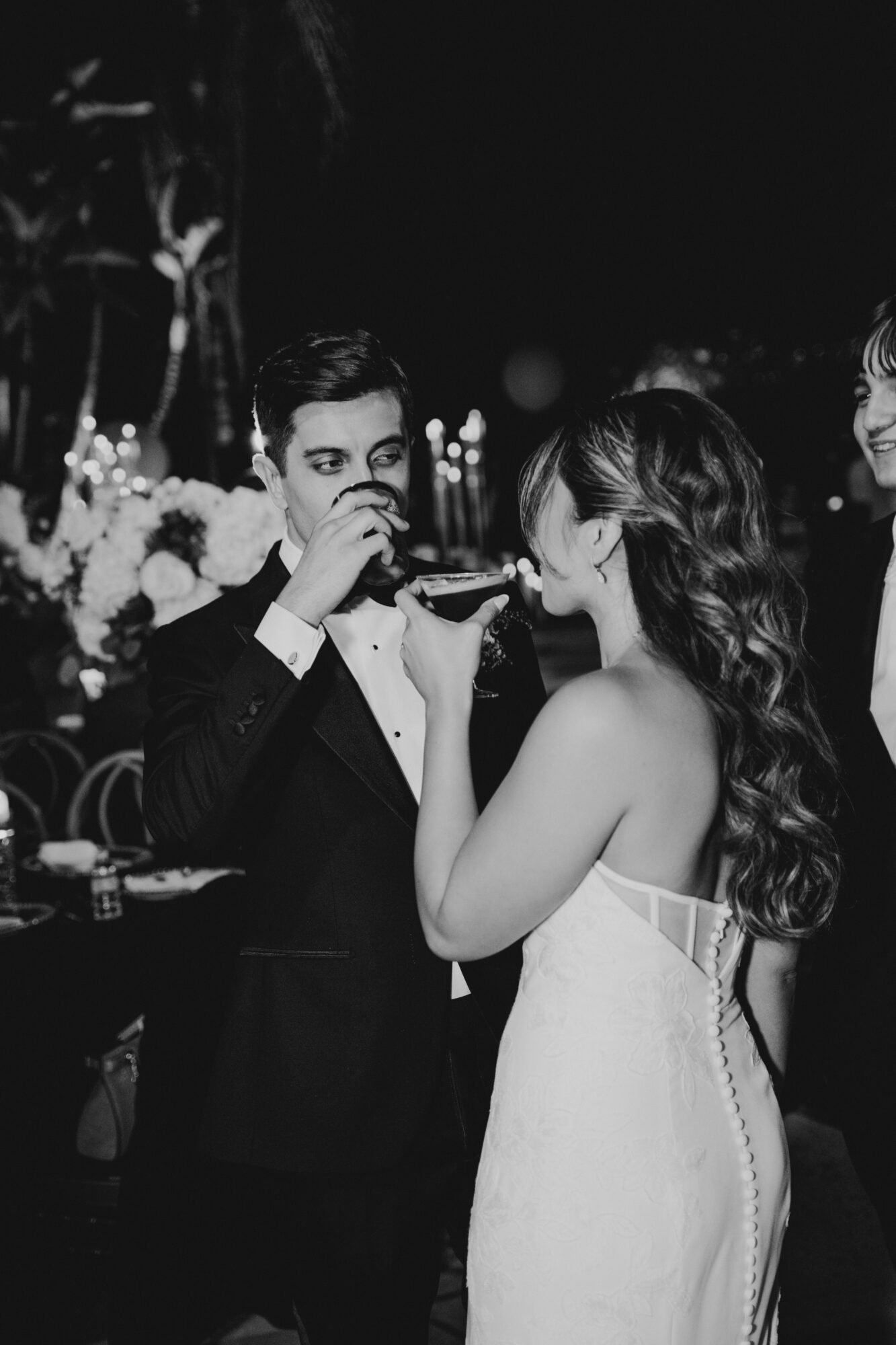 Man in tuxedo and woman in strapless dress sharing a drink at a formal event, with floral decorations in background.