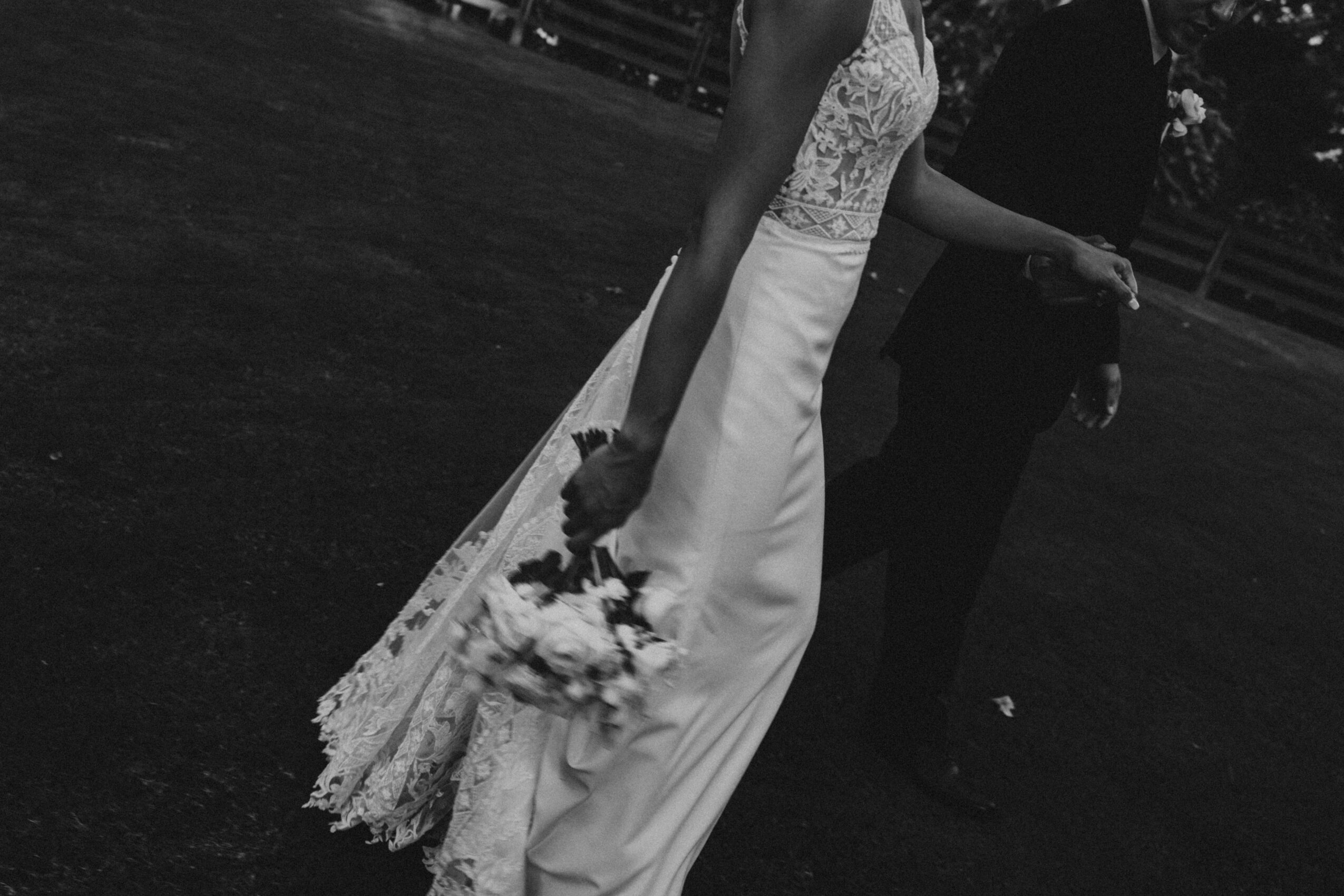 A woman in a wedding dress holding a bouquet, standing outdoors with a man in the background.