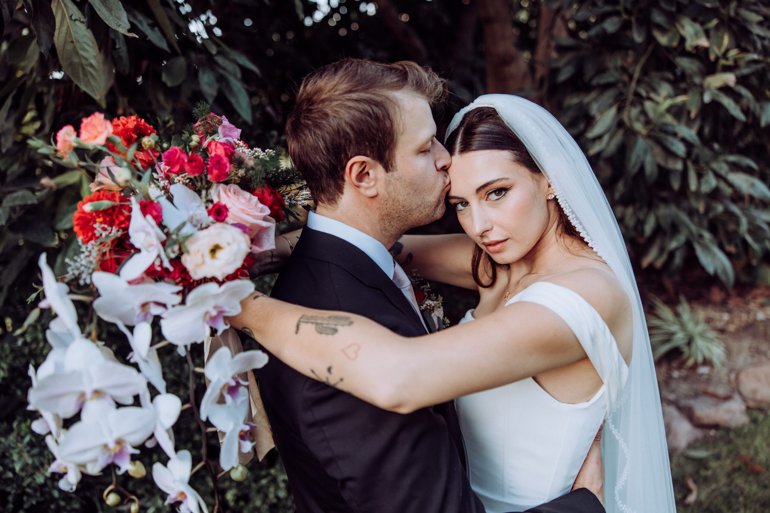 A bride and groom embrace outdoors, with a large bouquet of flowers nearby, surrounded by greenery.