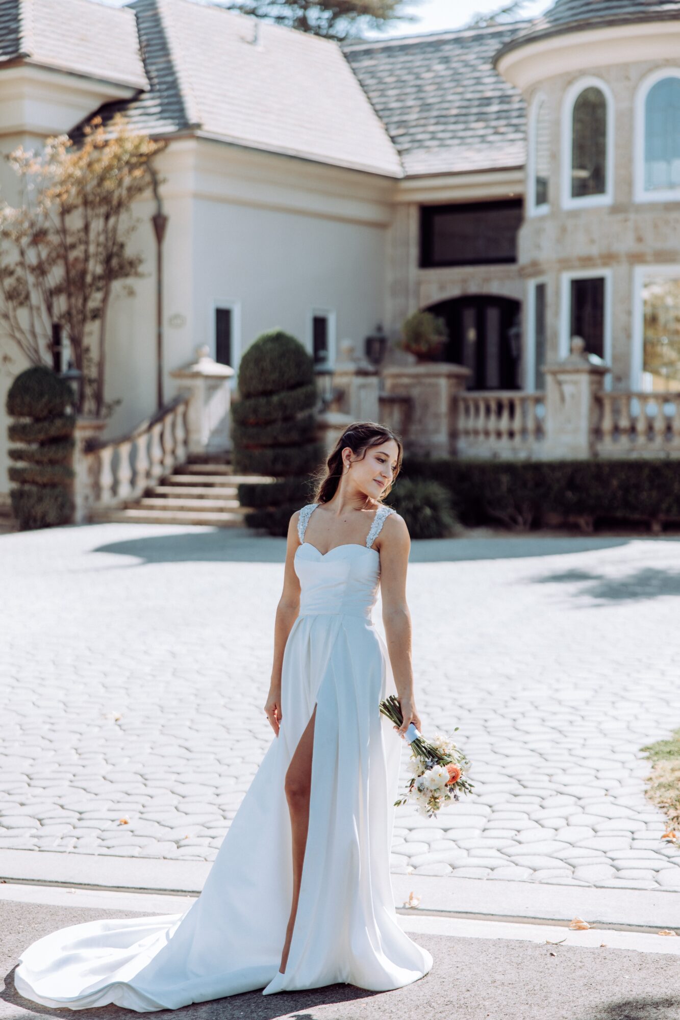 Young woman in a white wedding dress holding a bouquet, standing outdoors in front of a large house with a cobblestone driveway.