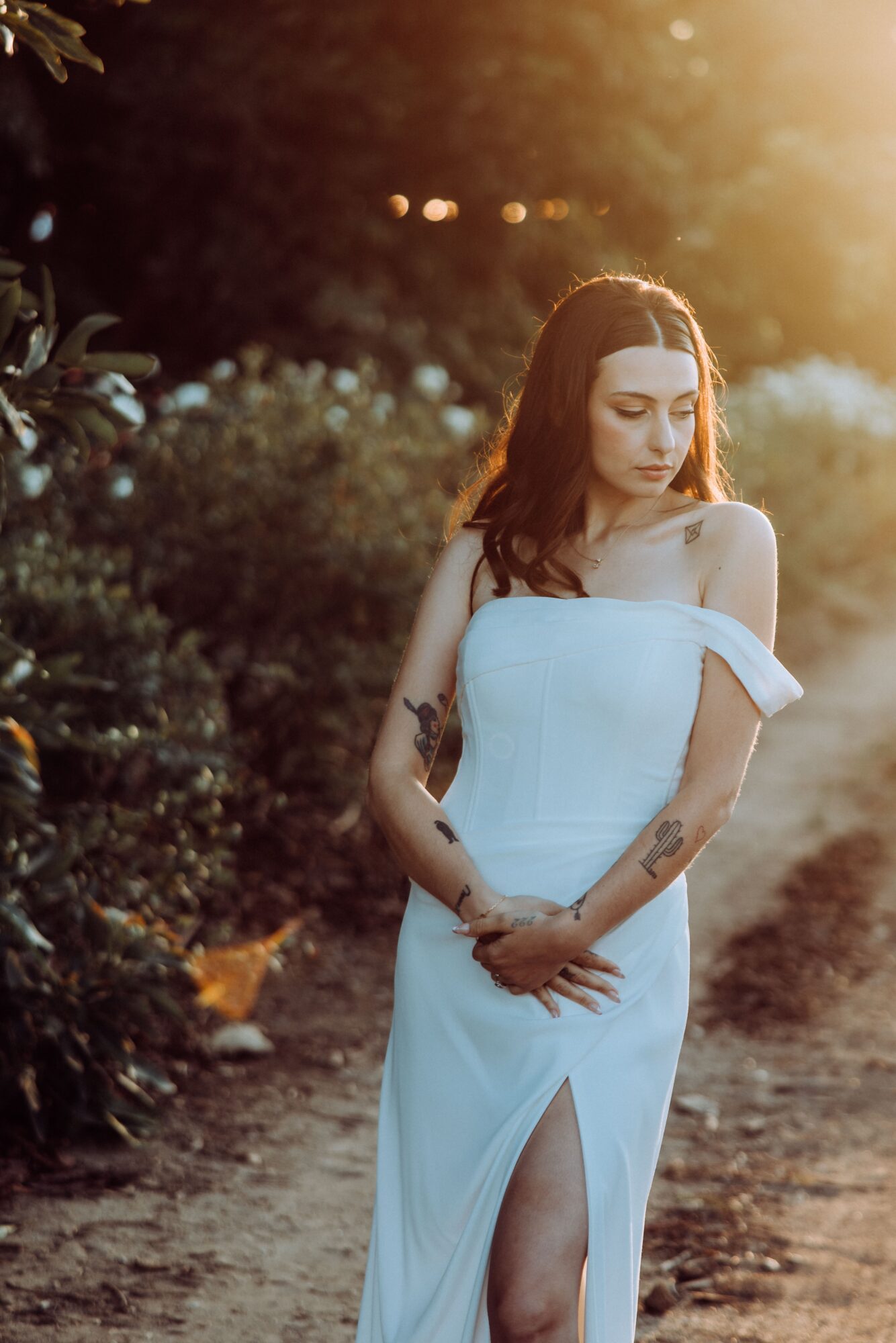 Young woman in white dress standing outdoors with sunlight, hands clasped, looking down, surrounded by greenery.
