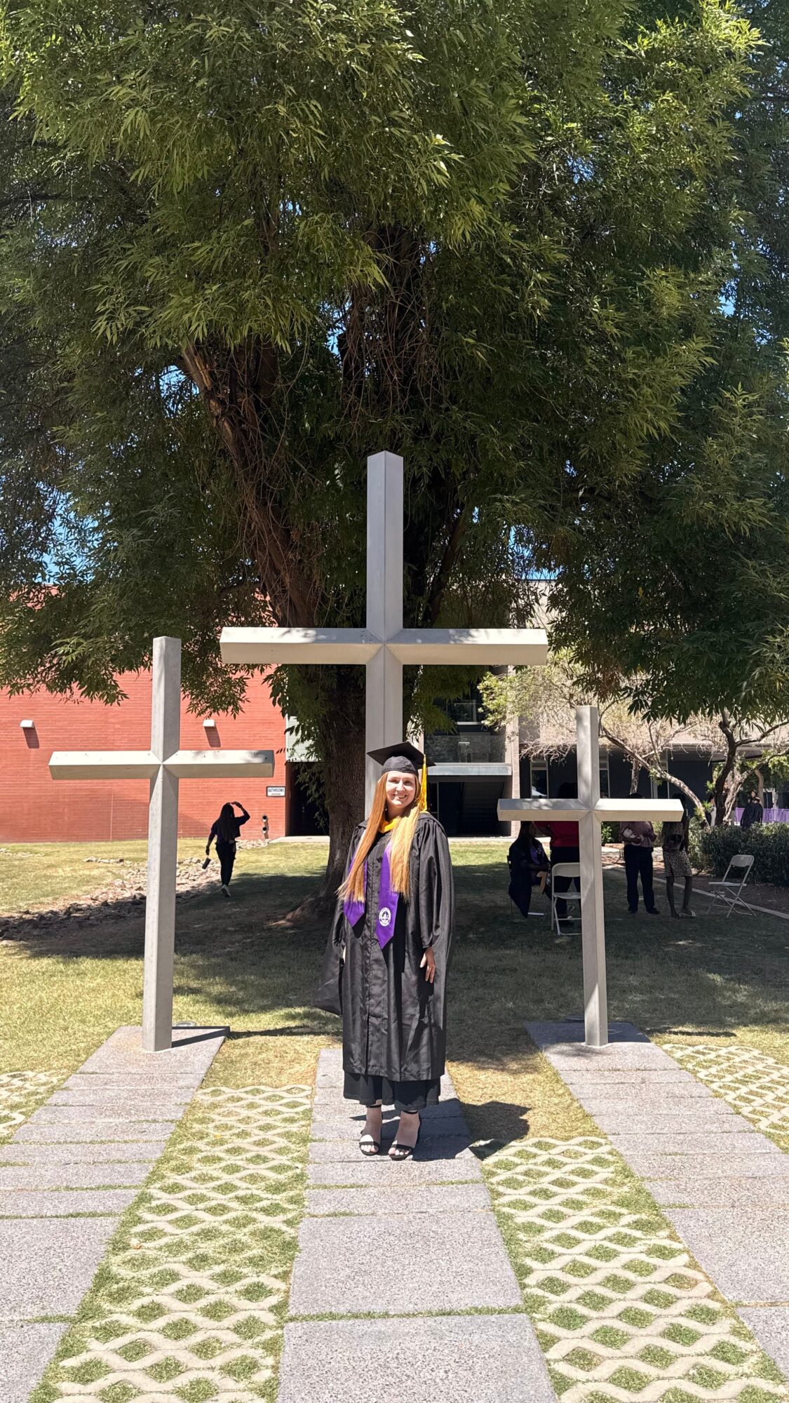 Person in graduation gown and cap standing under three large crosses outdoors, with trees and people in background.