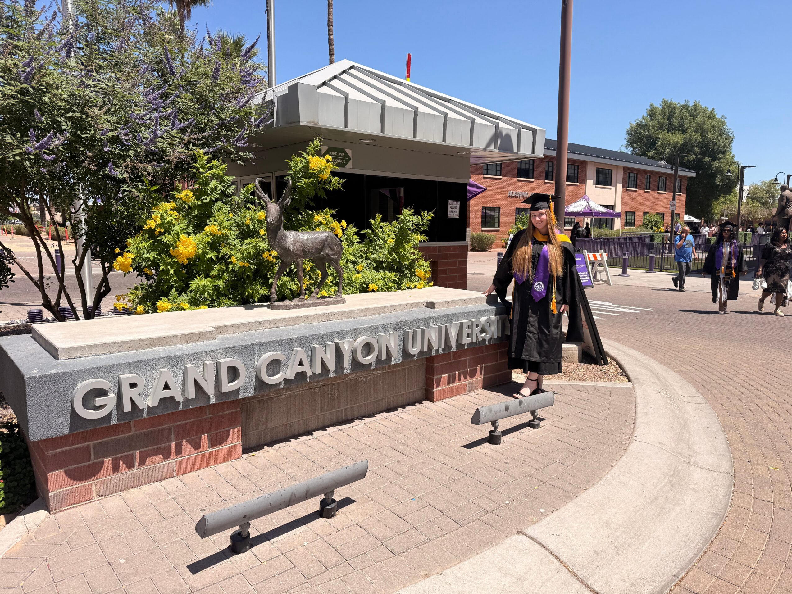 Person in graduation gown and cap standing near Grand Canyon University sign, with building and trees in background.