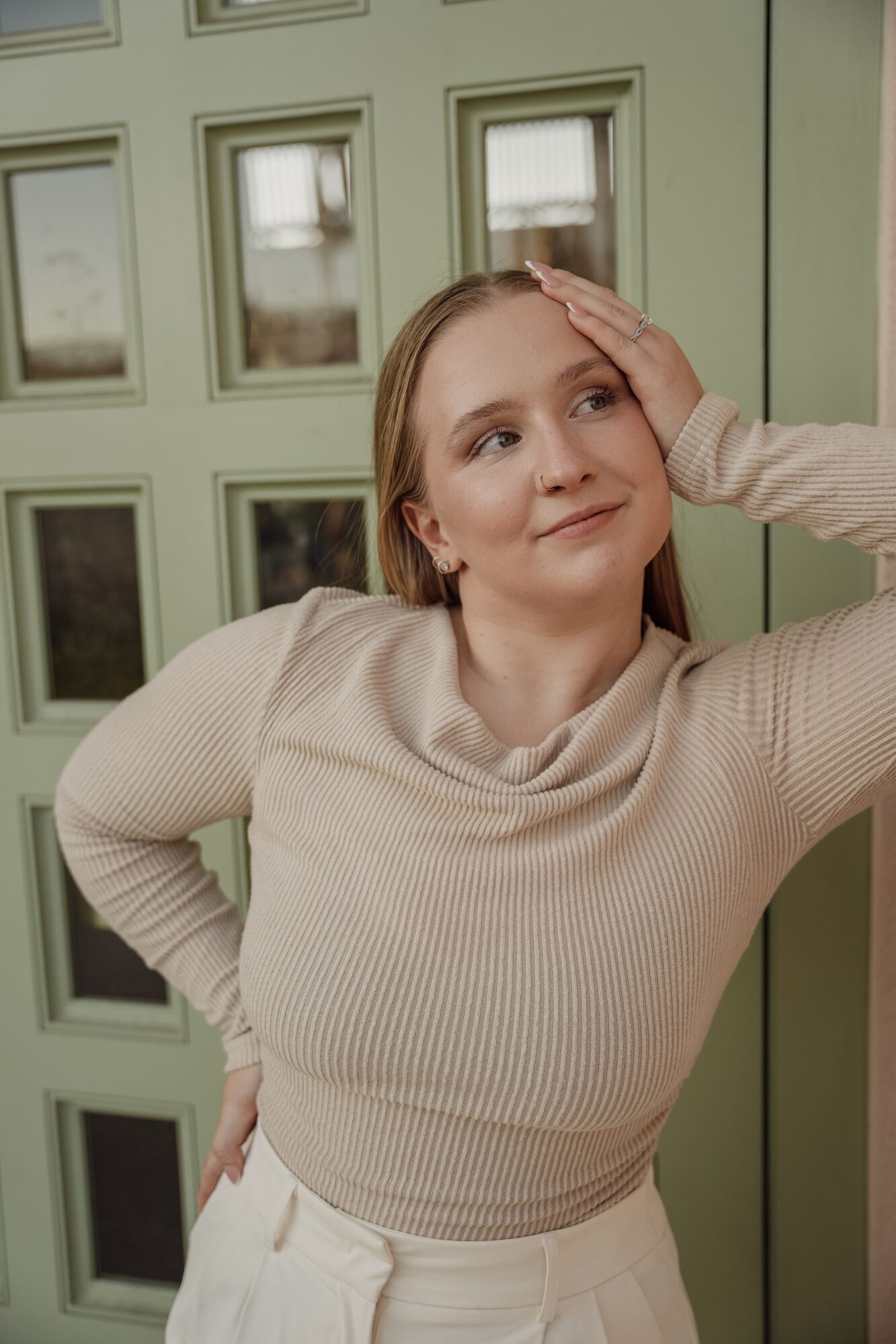 Woman with light brown hair leaning against a green door with glass panels, smiling gently, hand on forehead, wearing a beige sweater.