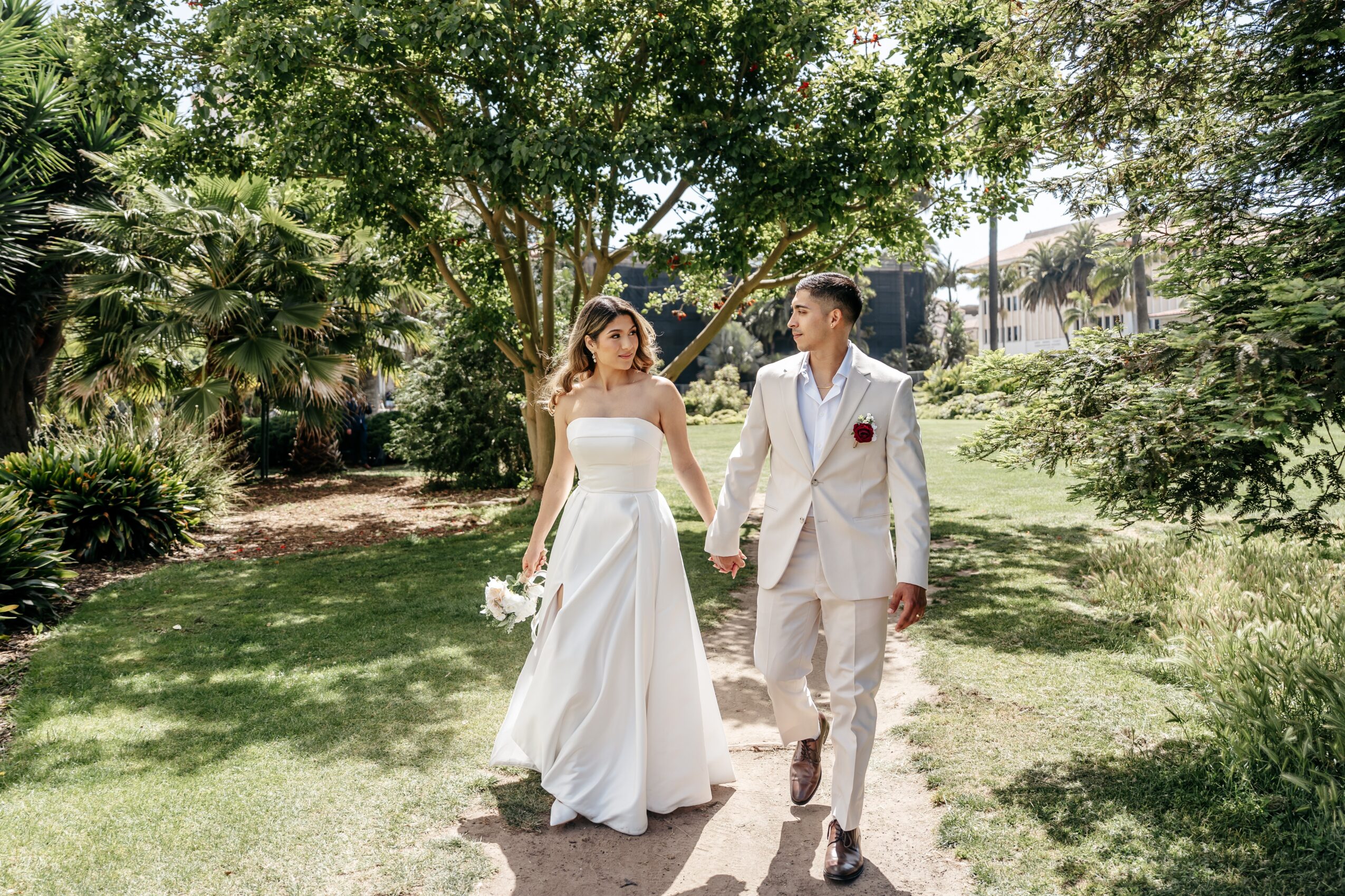 A bride and groom walk hand in hand in a park with trees and grass, holding flowers, dressed in wedding attire.