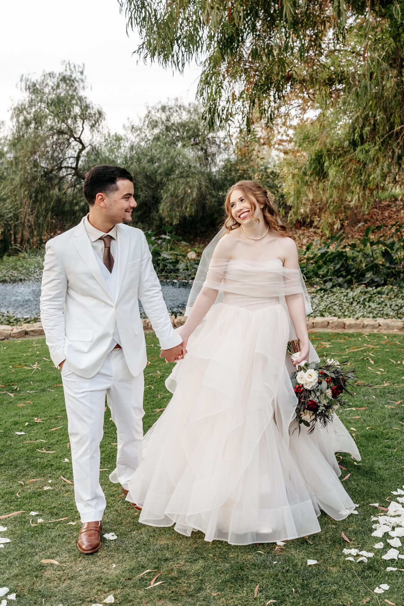 A bride and groom holding hands outdoors, smiling, with trees and a river in the background.