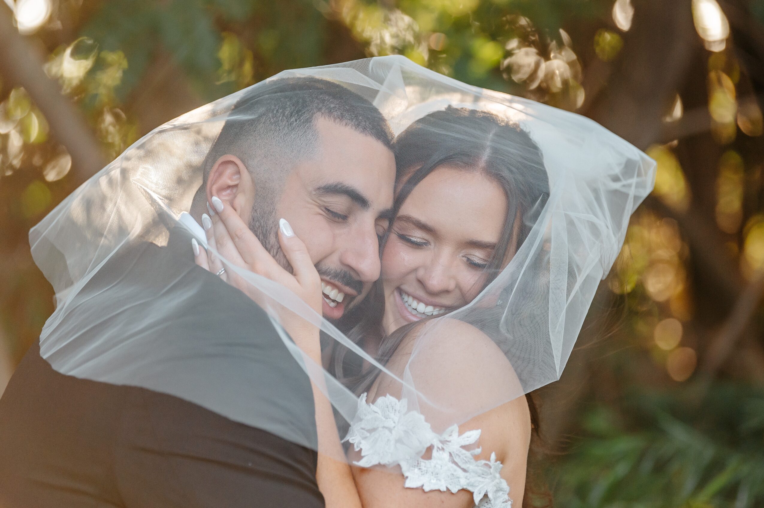 A bride and groom smiling and embracing under a veil outdoors, with trees in the background.