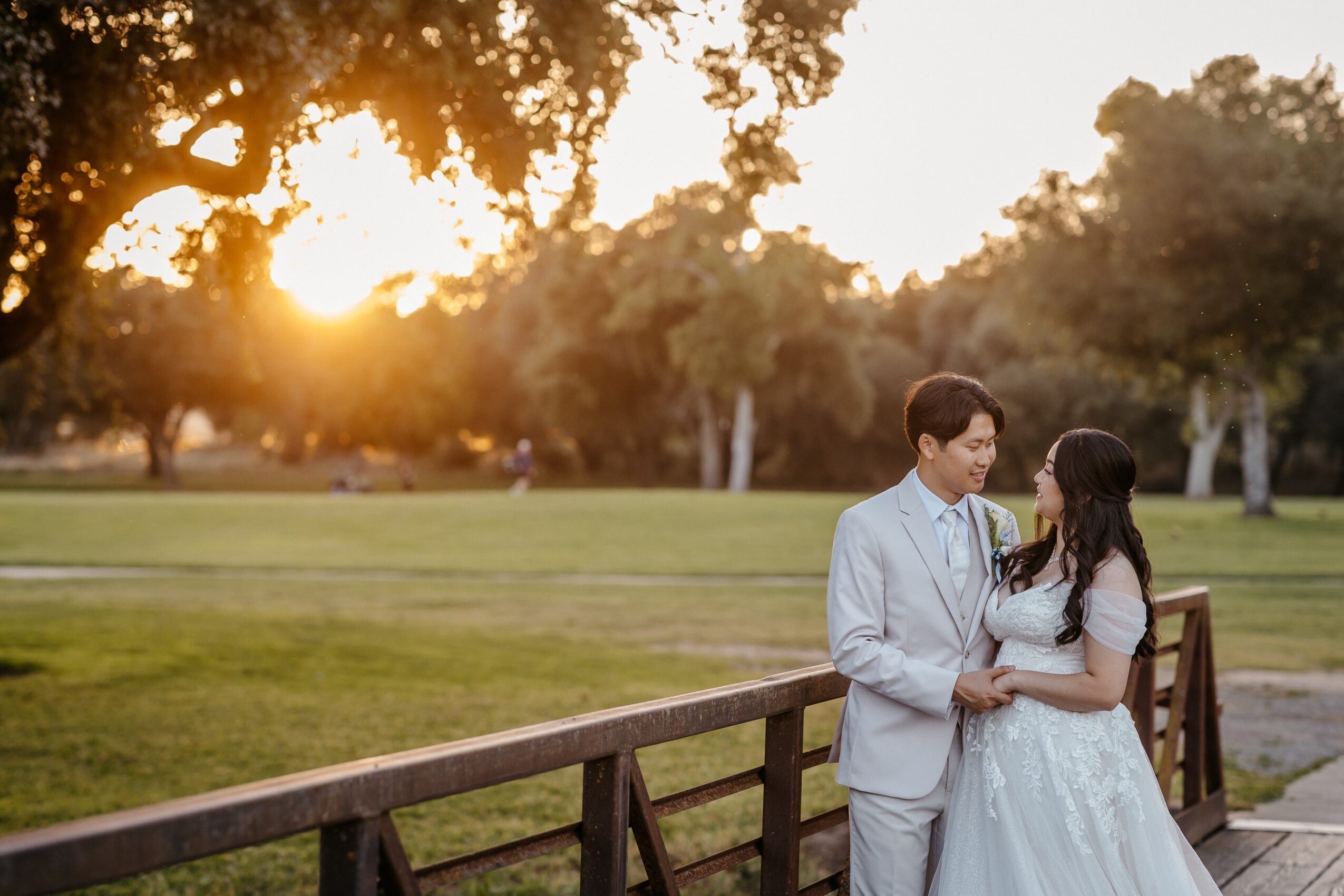 A couple stands on a wooden bridge in a park during sunset, holding each other and looking at each other.