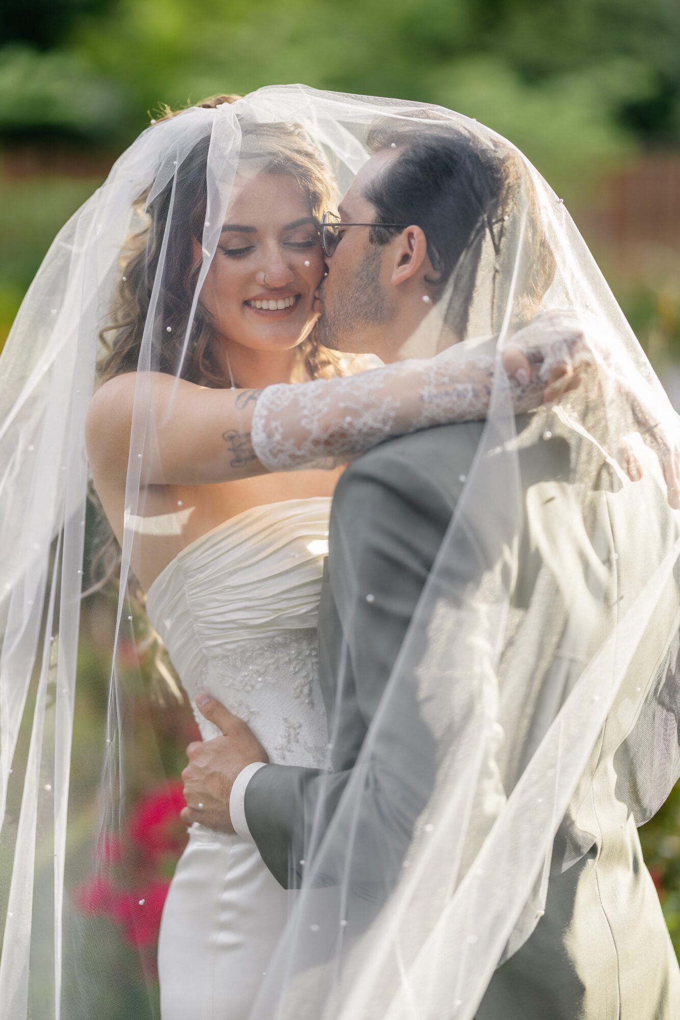 Bride and groom smiling under a veil outdoors, embracing each other, with greenery in the background.