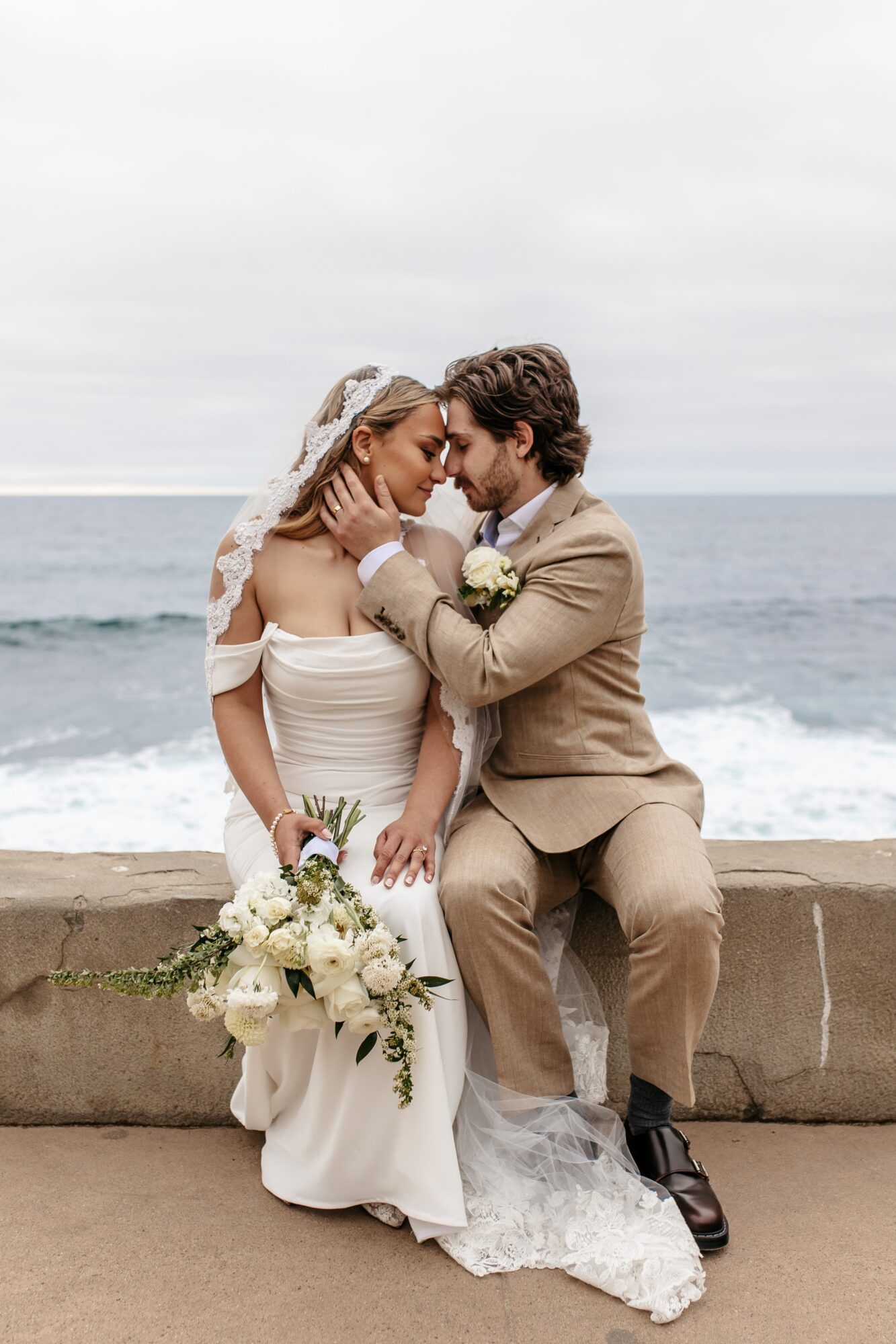 Bride and groom sit close on a wall by the sea, holding hands and gazing at each other, with wedding attire and bouquet.