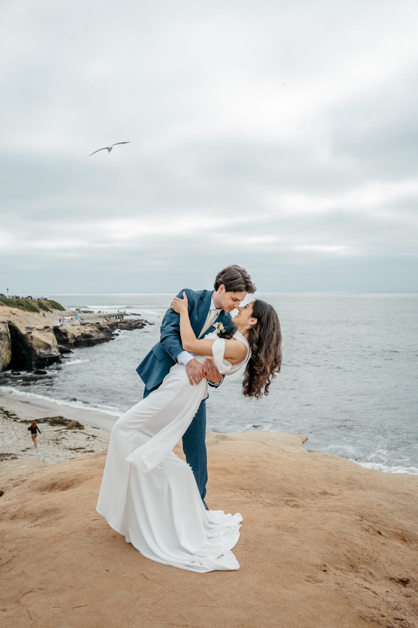 A couple dancing on a beach with rocks and ocean in the background, overcast sky, one person in a white dress, the other in dark clothing.