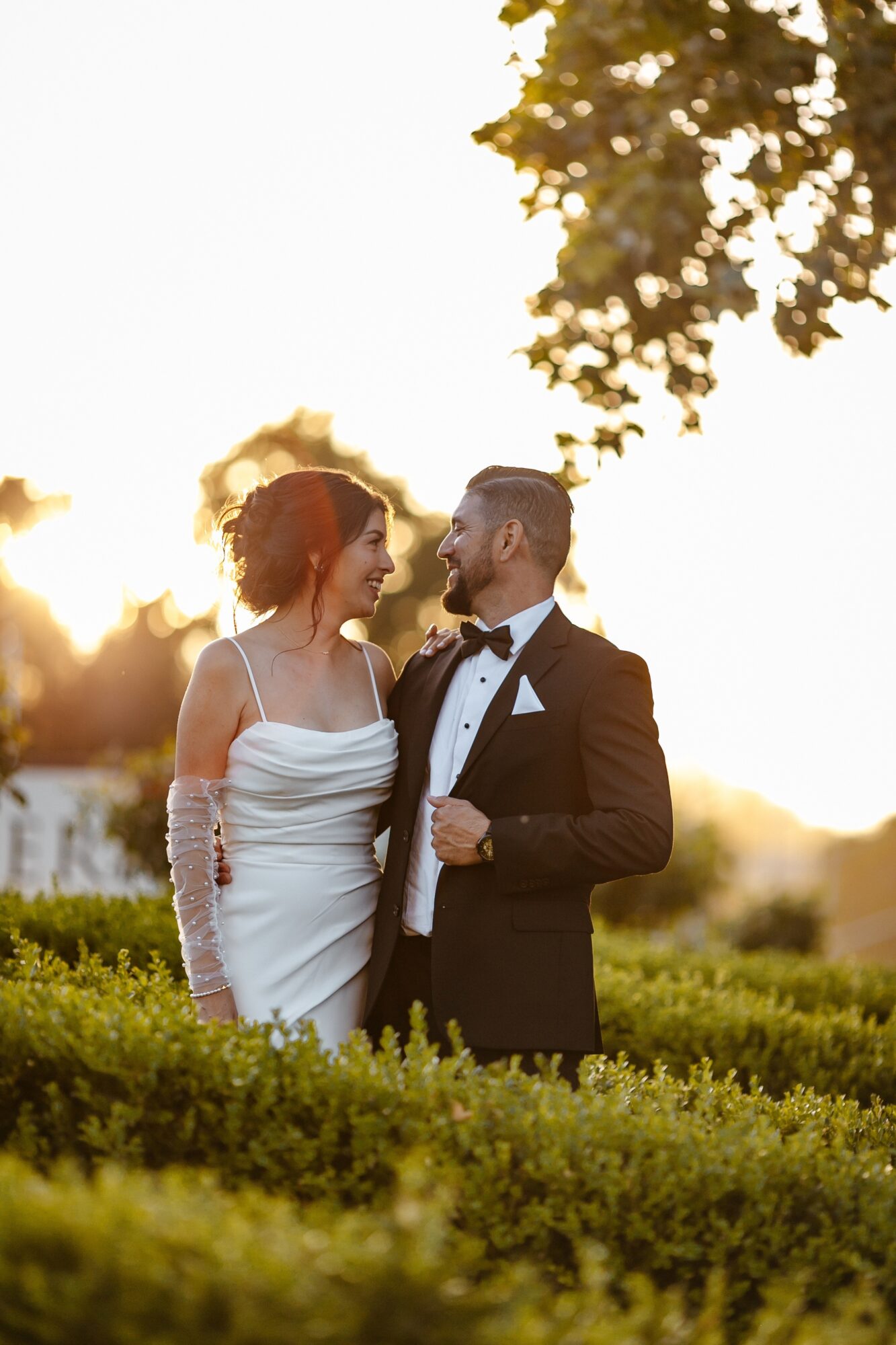 A couple in wedding attire standing outdoors with trees and sunlight in the background.