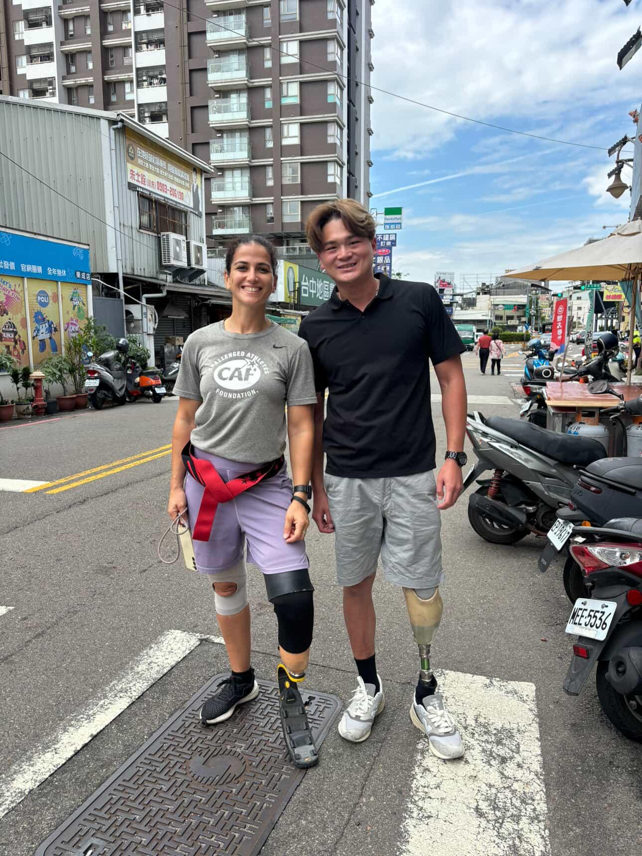 Two people standing on a city street, smiling, with buildings and parked scooters in the background.