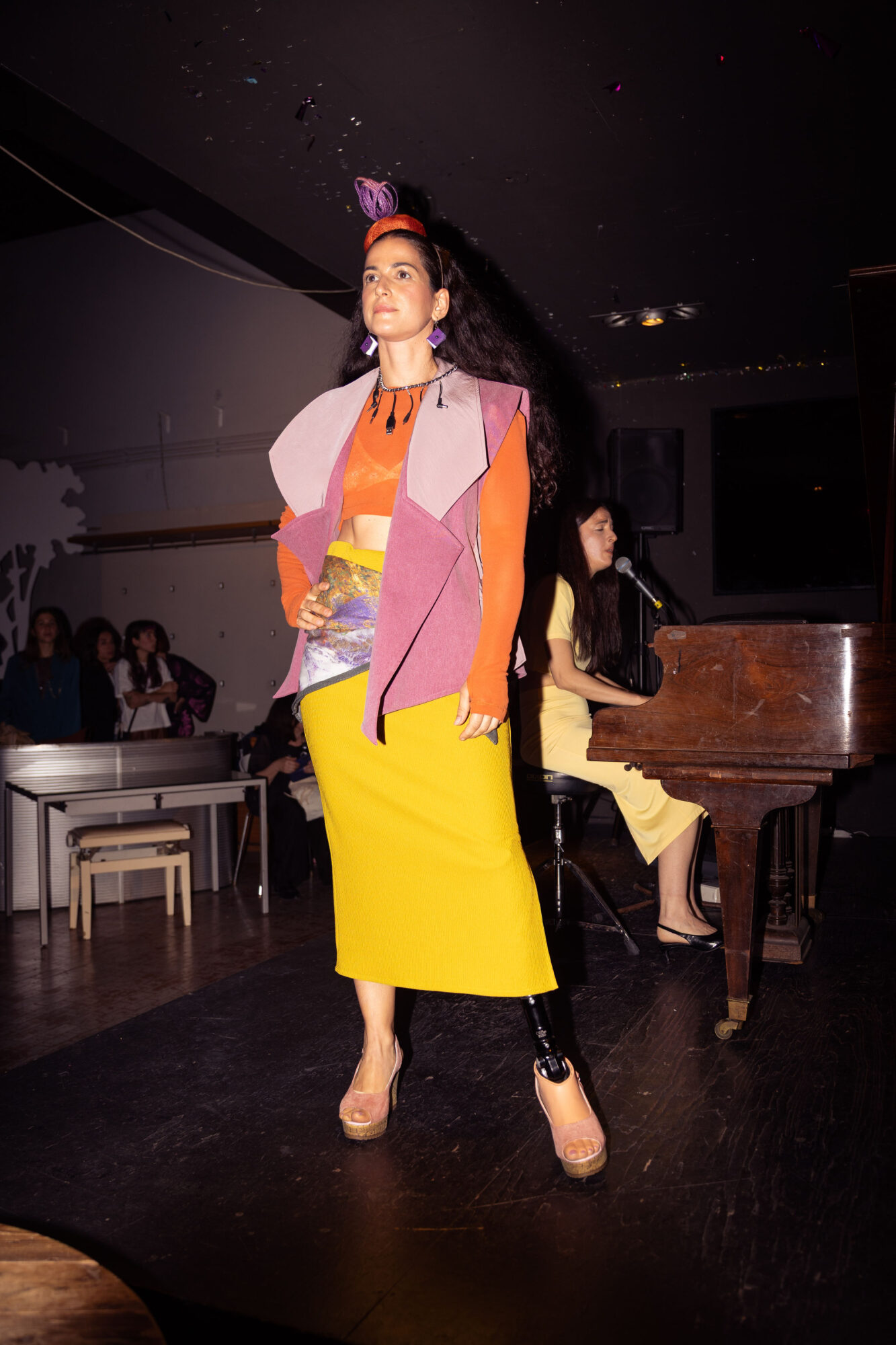 Woman standing on stage in colorful outfit, with a person playing piano in background, audience seated nearby.