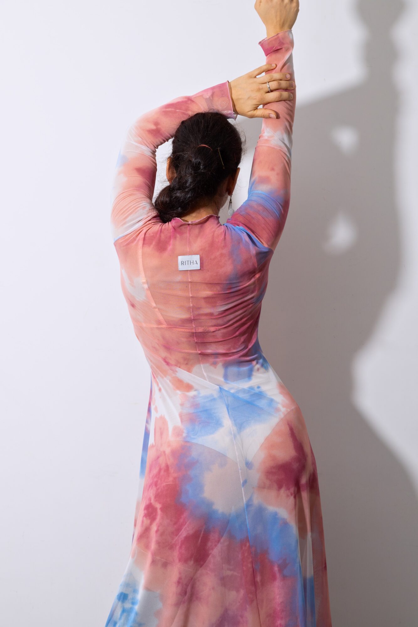Person with dark hair in a colorful, tie-dye dress stretching arms upward, facing away from the camera, with a shadow on the wall.