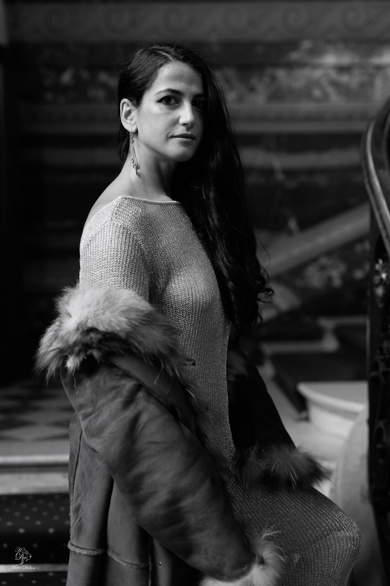 Woman with long dark hair wearing a sweater and fur coat, standing indoors with blurred background.
