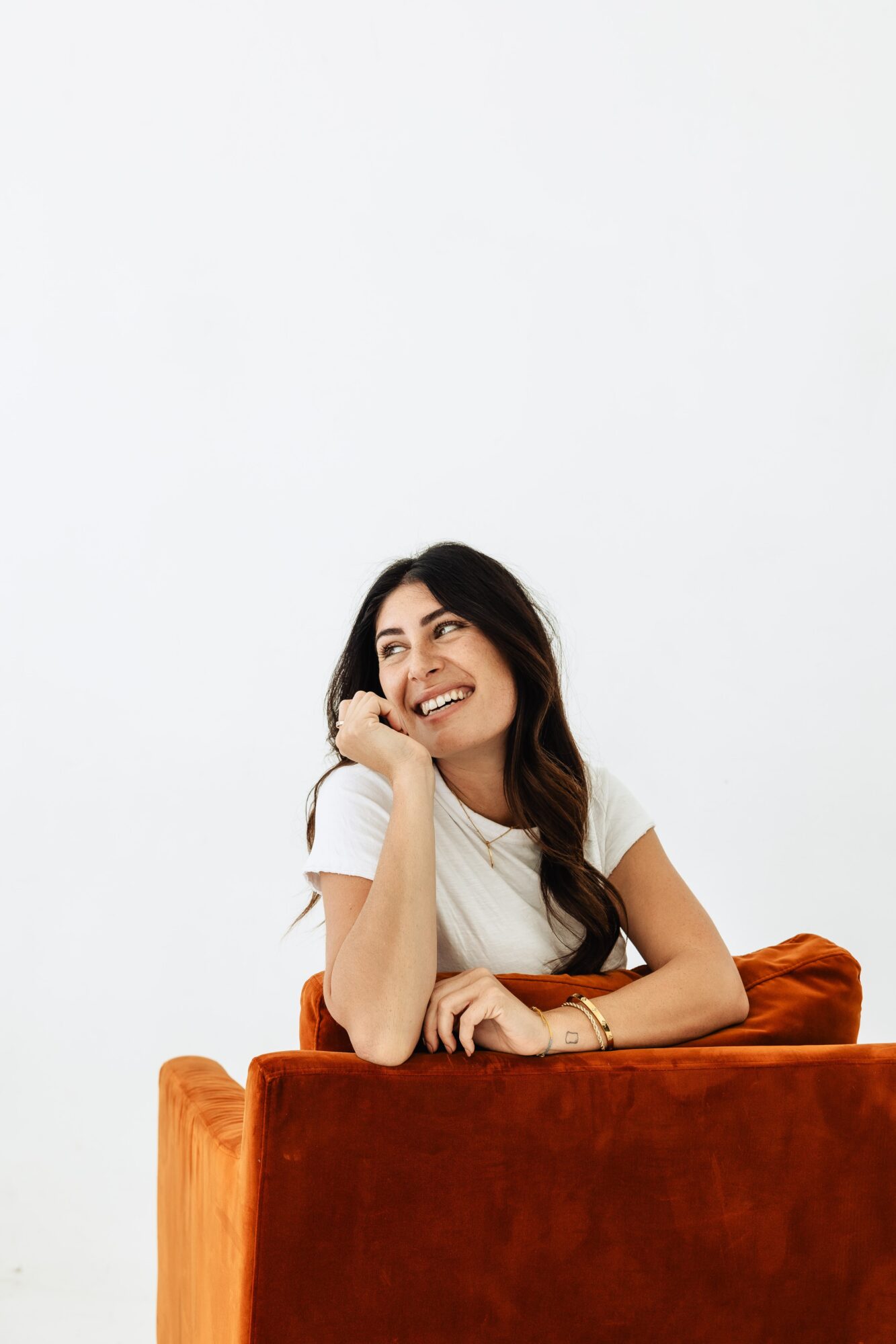 Young woman with long dark hair smiling, resting her chin on her hand, sitting behind a rust-colored sofa, white background.