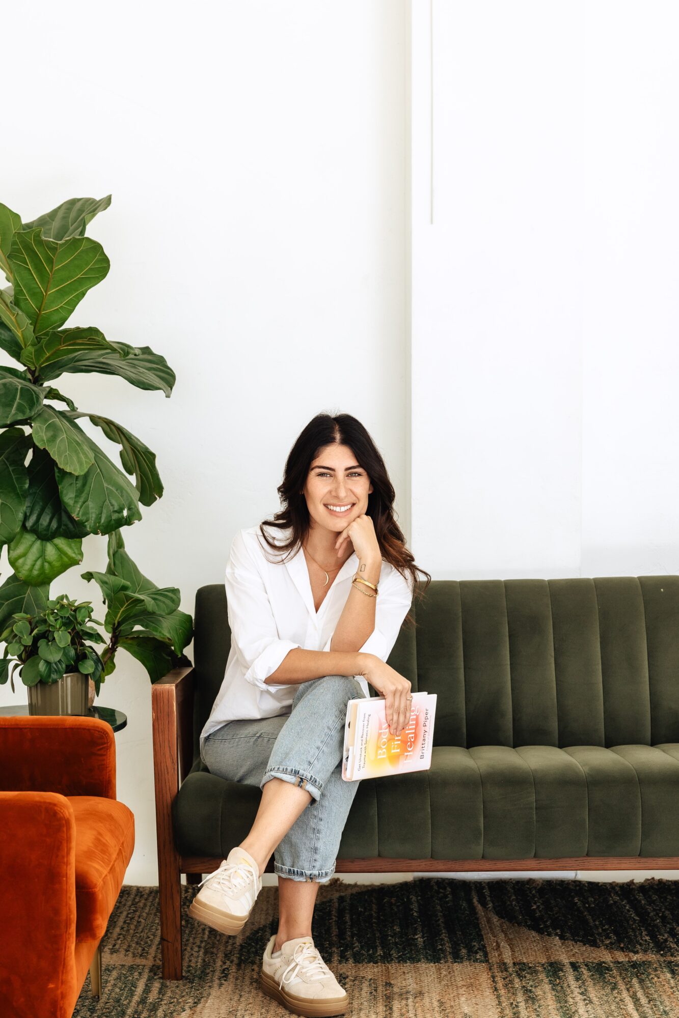 Woman sitting on a green sofa, smiling, holding a colorful book, in a room with a large plant and white wall.