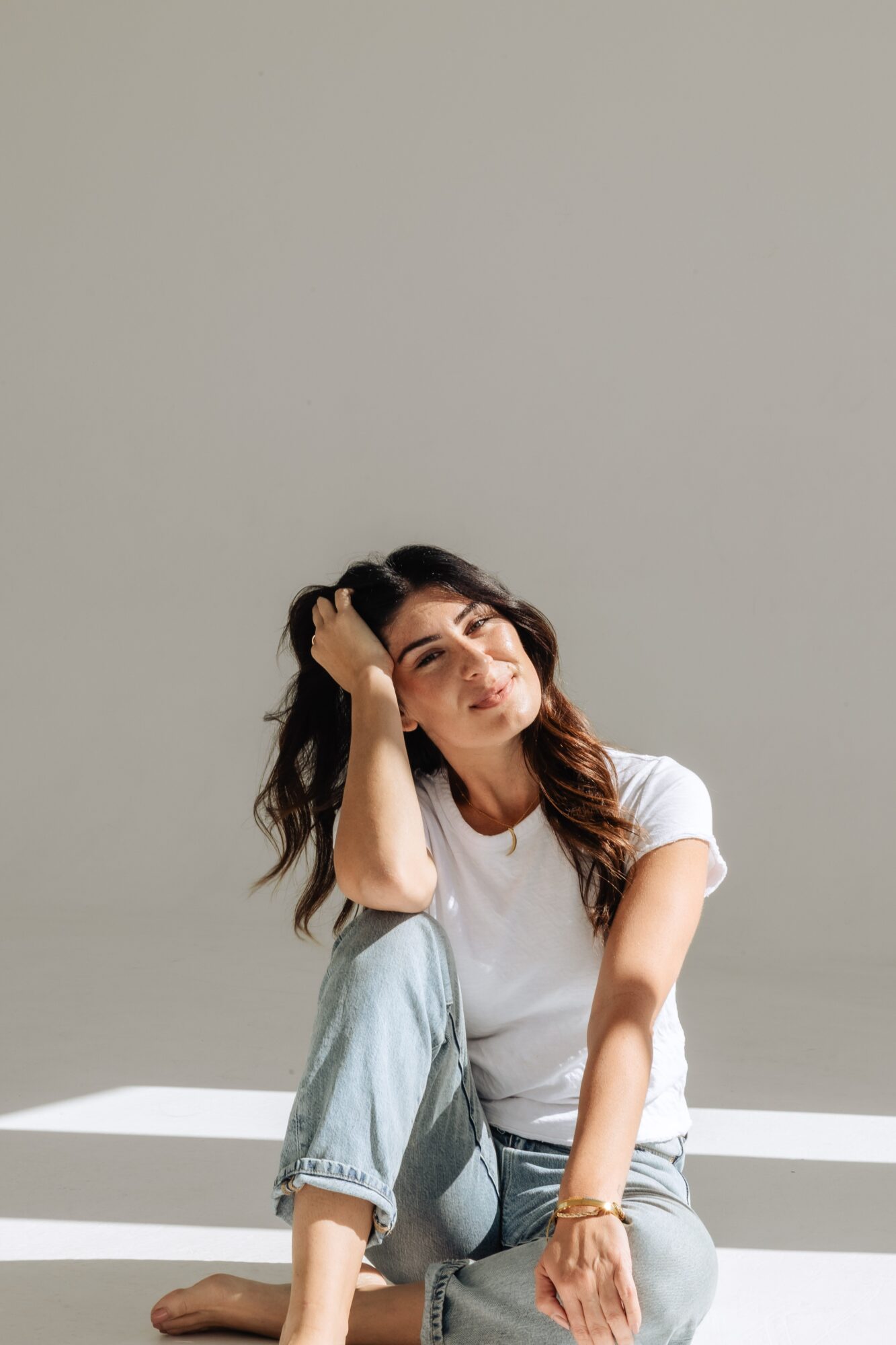 Woman sitting on a white surface, resting her head on her hand, wearing a white t-shirt and light blue jeans.